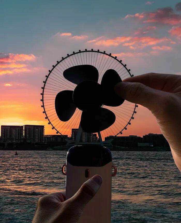 A hand holding a fan in front of a Ferris wheel, a clever photo manipulation making simple objects look like parts of cities.