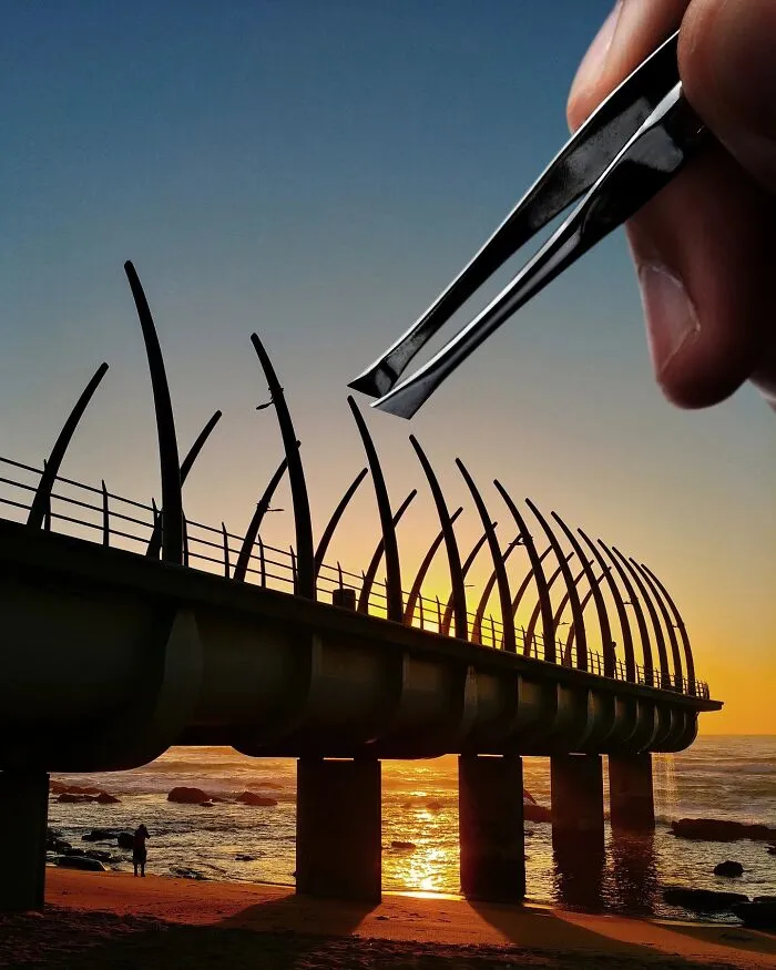 A hand holding tweezers above a pier at sunset. This photo manipulation makes simple objects look like parts of cities.