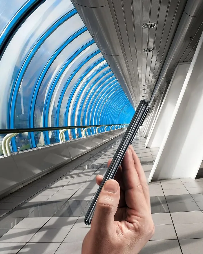 A hand holding a phone, aligning it with a modern blue and white tunnel. This photo manipulation makes simple objects look like parts of cities.