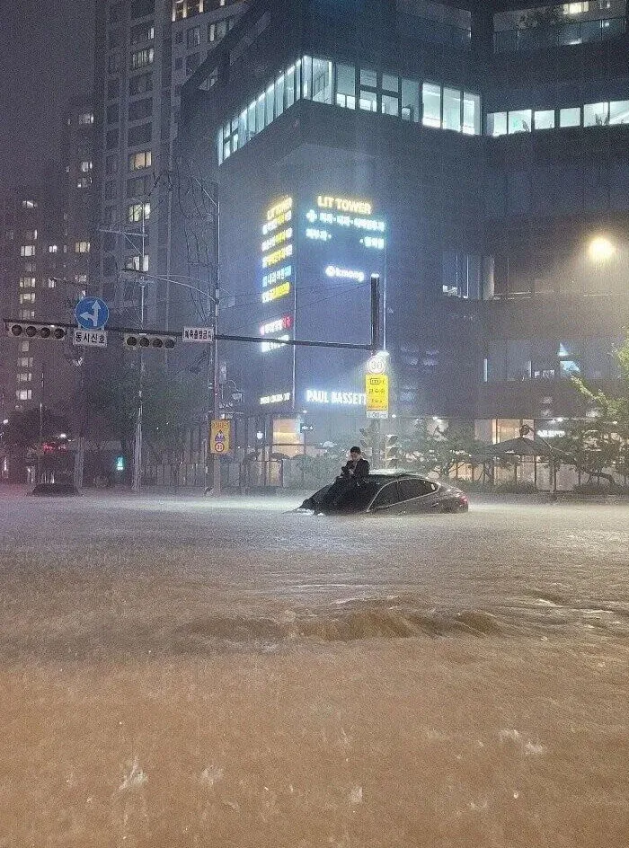 Flooded urban street with submerged car and person sitting on top during heavy rain