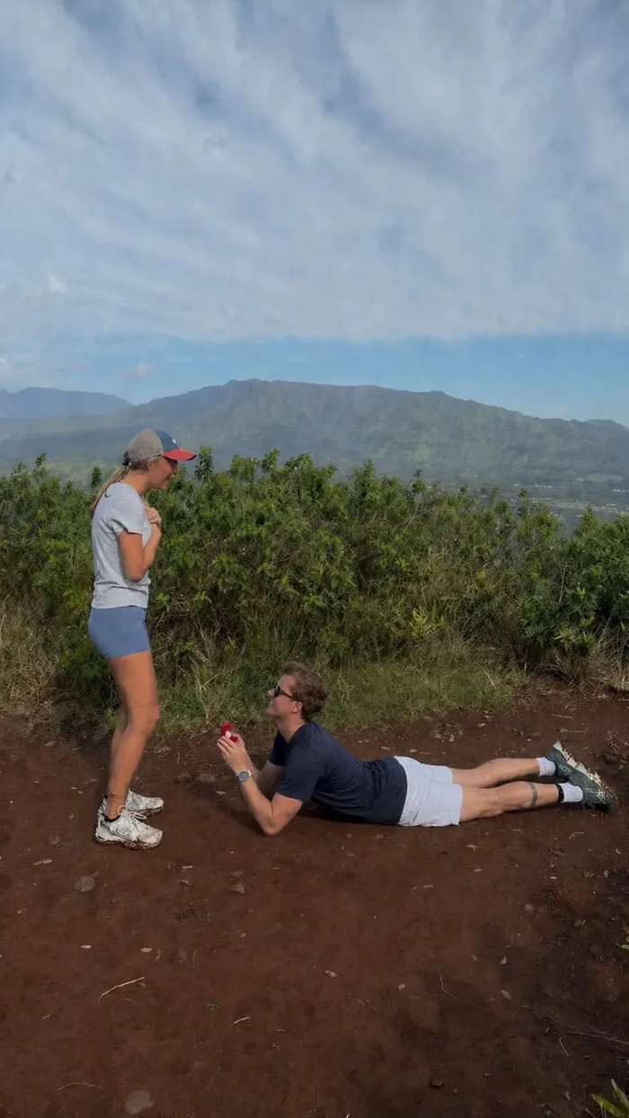 Woman reacting to a man's proposal pic, lying down with a ring box, on a hiking trail overlooking mountains.