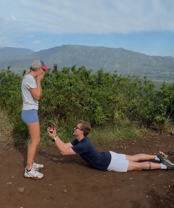 Man proposing to woman in a memorable proposal pic, lying on the ground, holding a ring box, with mountains in background.