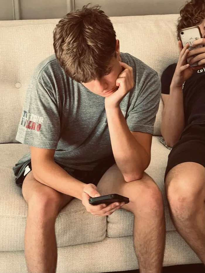 A young man with brown hair intently looks at his phone while another holds their phone. This is typical Gen Z behavior.