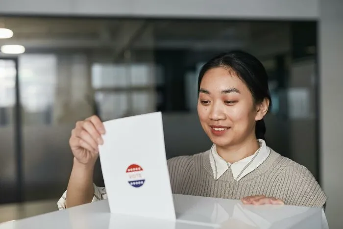 A young Asian woman, a Gen Z, smiles as she inserts a ballot with a "VOTE" sticker into a box.