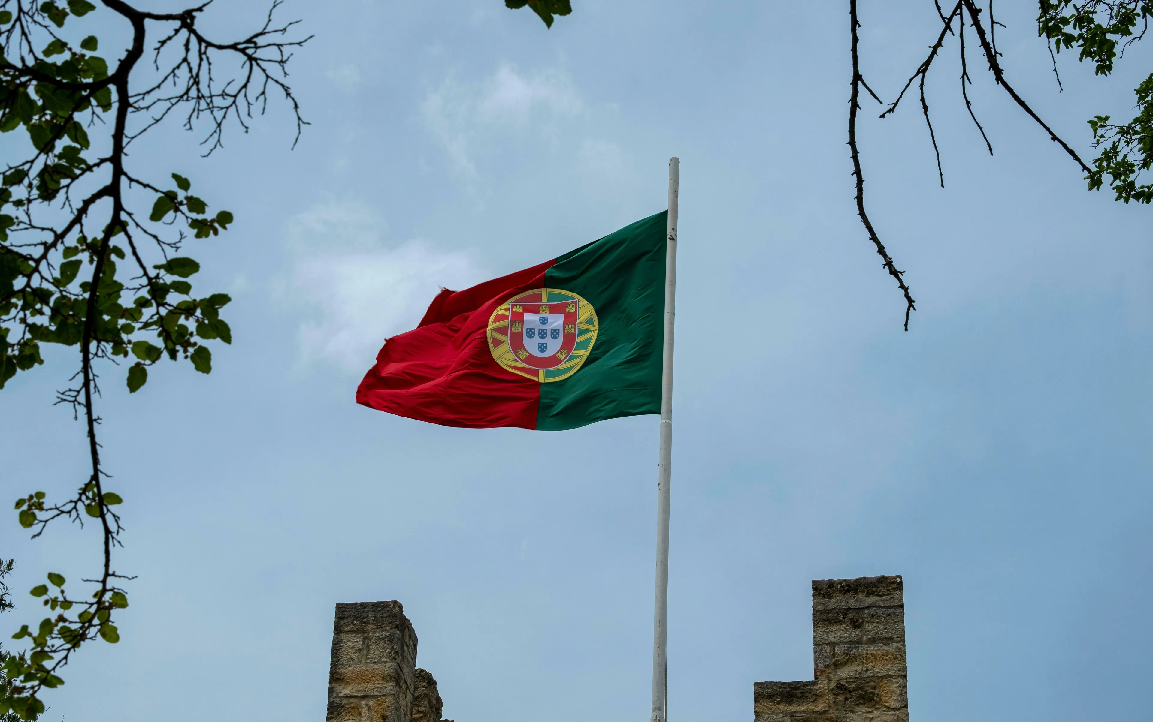 General knowledge quiz: Portugal's flag flying against a cloudy sky, framed by tree branches and old stone walls.