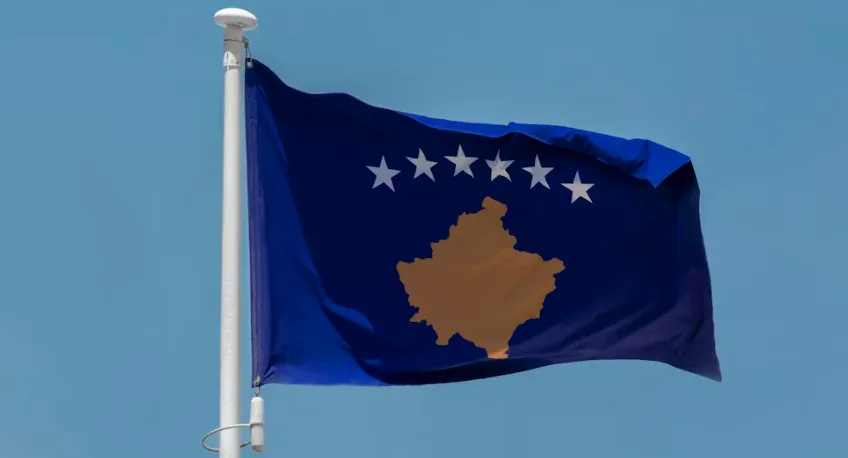 The flag of Kosovo, blue with a gold map and six white stars, against a clear blue sky. General Knowledge Quiz: Europe.