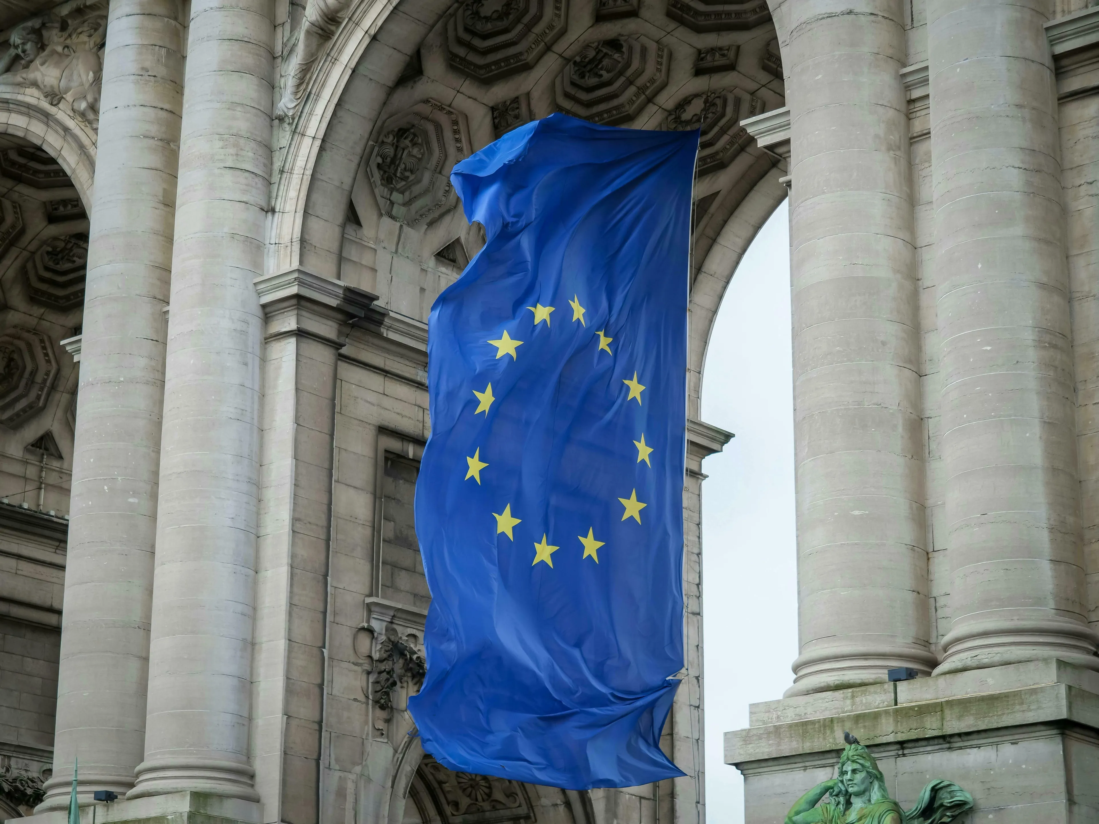 A European Union flag hangs from a grand stone archway, representing the de facto capital of the European Union.