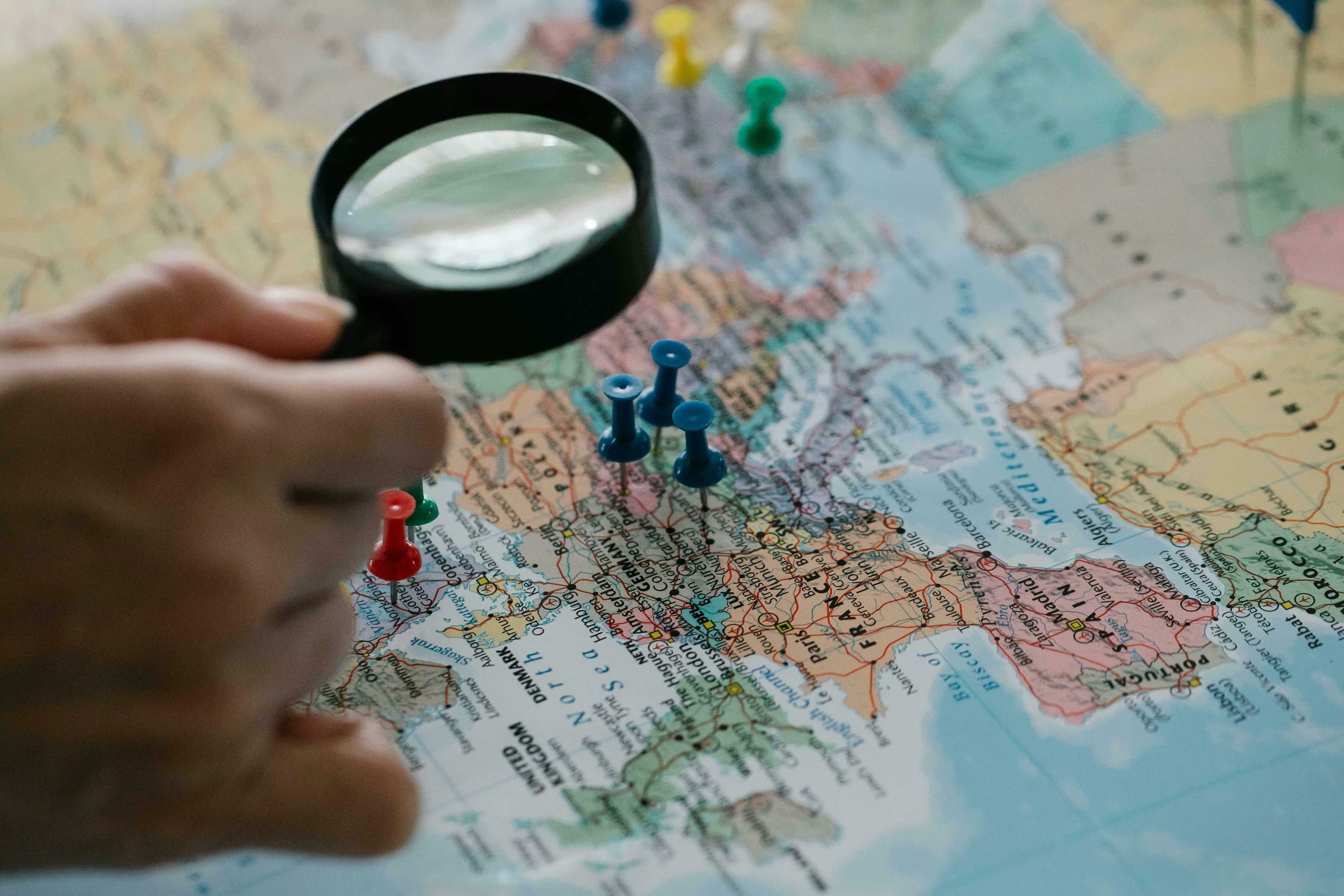 Hand using a magnifying glass on a Europe map with pushpins. General knowledge quiz about Europe questions.