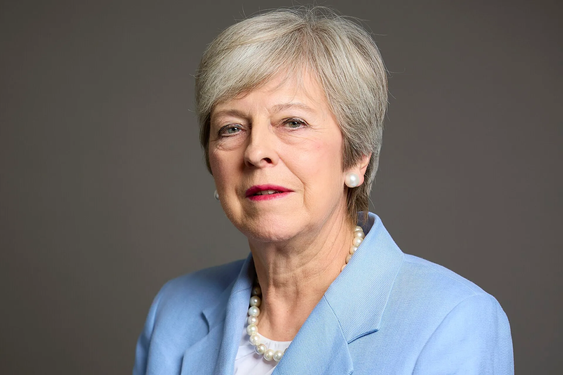 Close-up of a British former Prime Minister, wearing a blue blazer and pearl necklace, against a gray background.