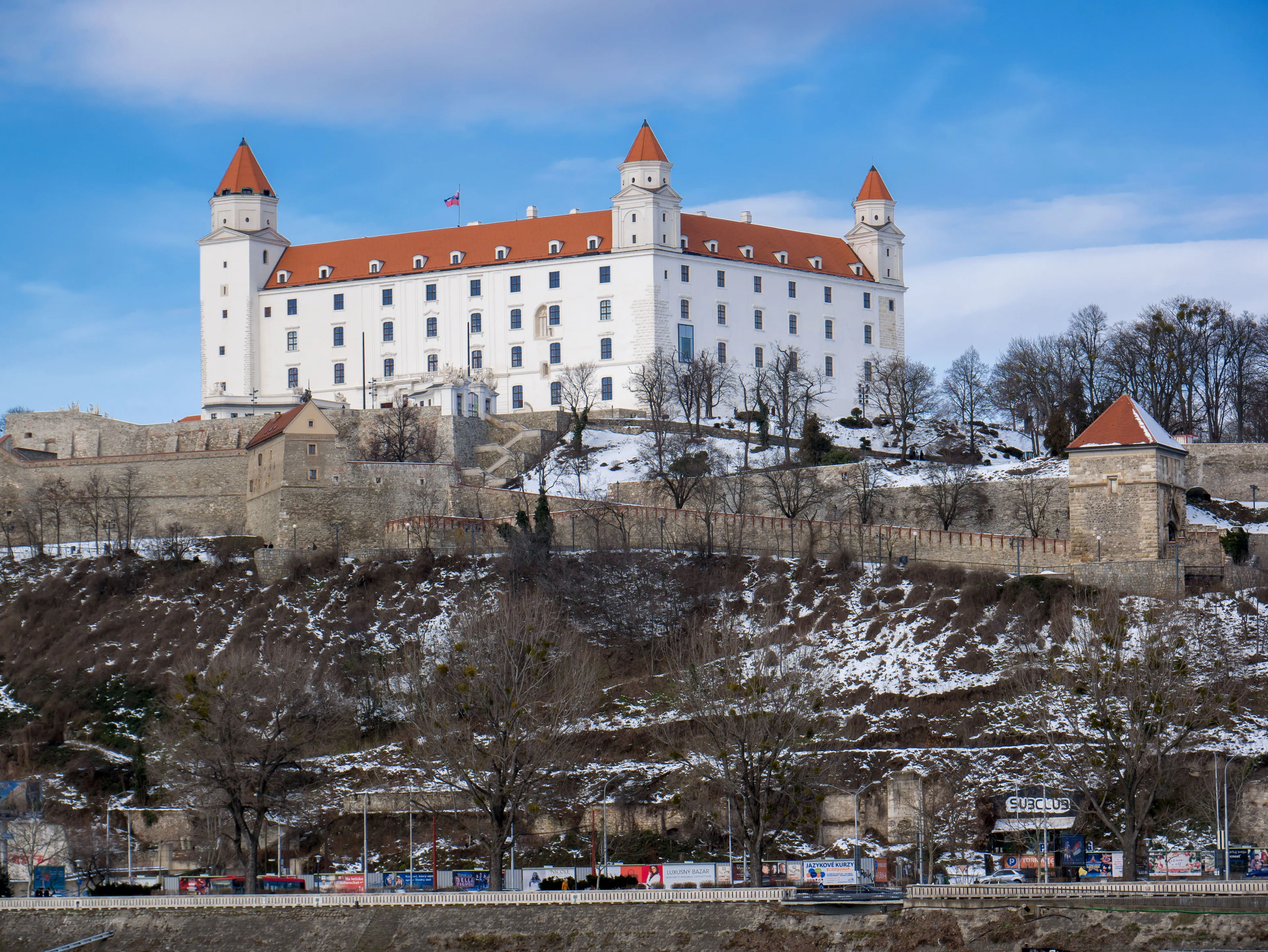 Bratislava Castle, a grand white structure with red roofs, perched on a snowy hill, overlooking the capital.