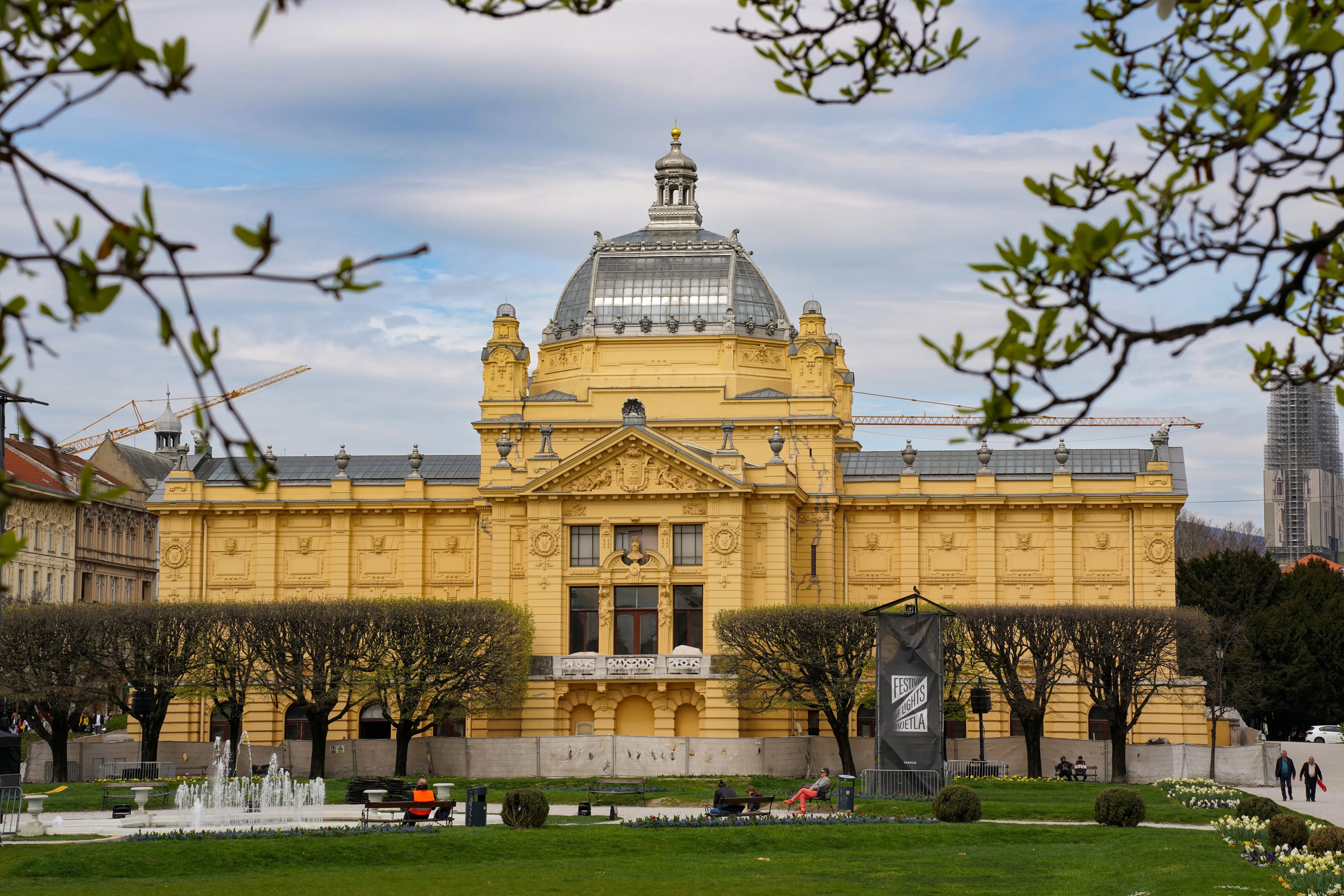 A grand yellow building with a dome, surrounded by green grass and trees in Zagreb, the capital of Montenegro.