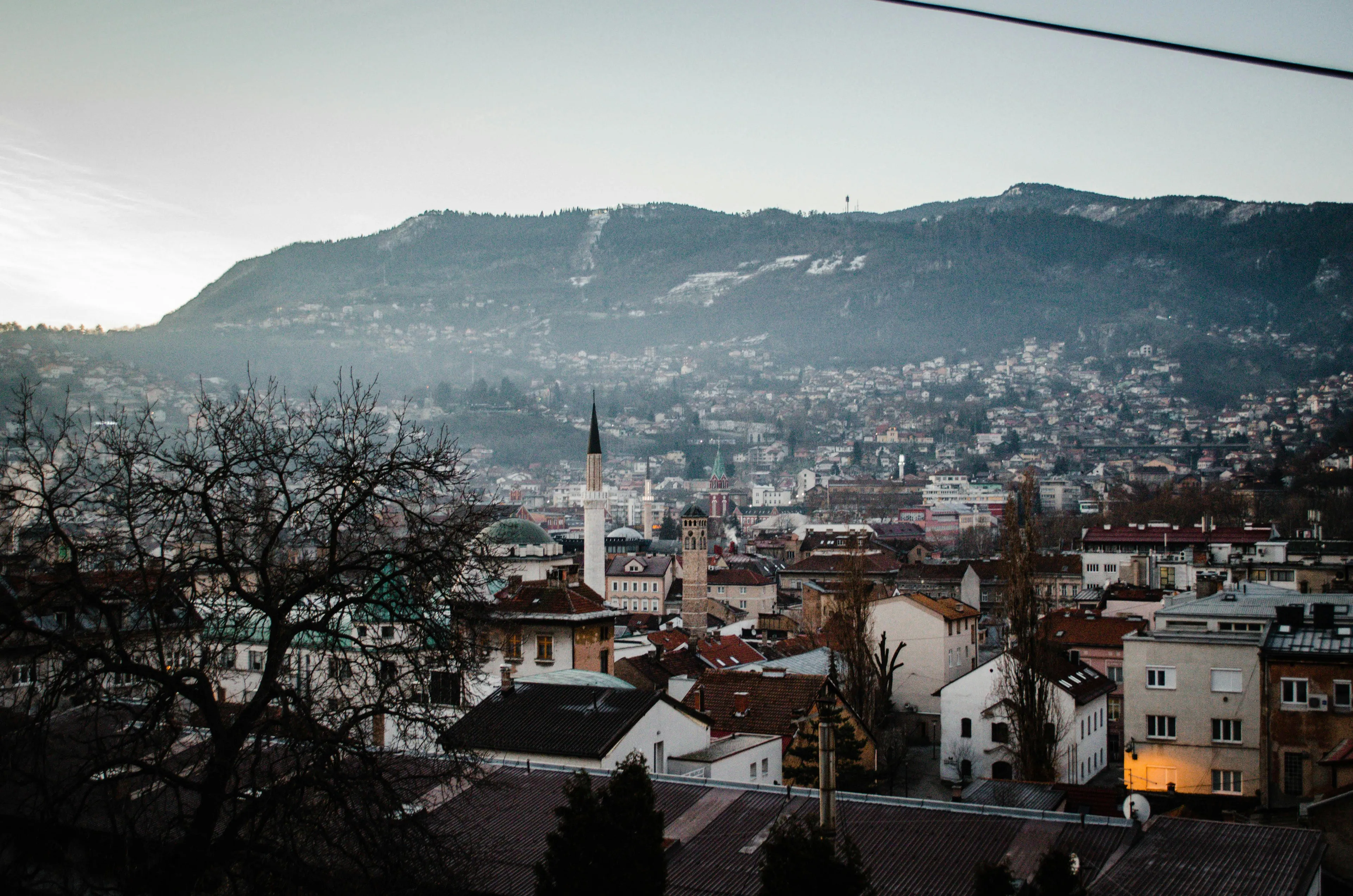 Sarajevo cityscape with a mosque minaret and historical buildings under a cloudy sky, surrounded by hills.