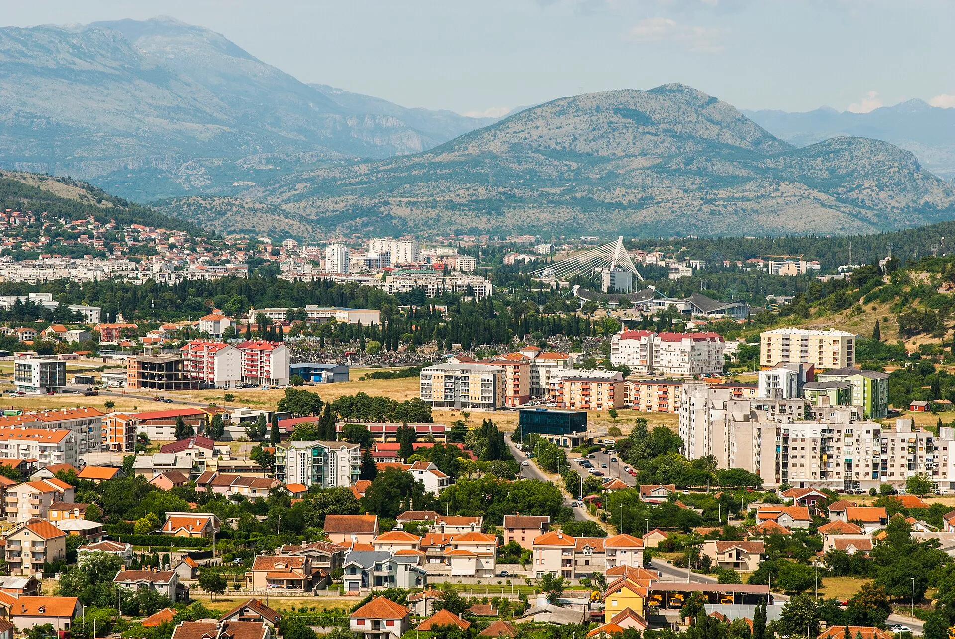 A panoramic view of Podgorica, the capital of Montenegro, featuring urban buildings nestled among green trees with mountains in the background.