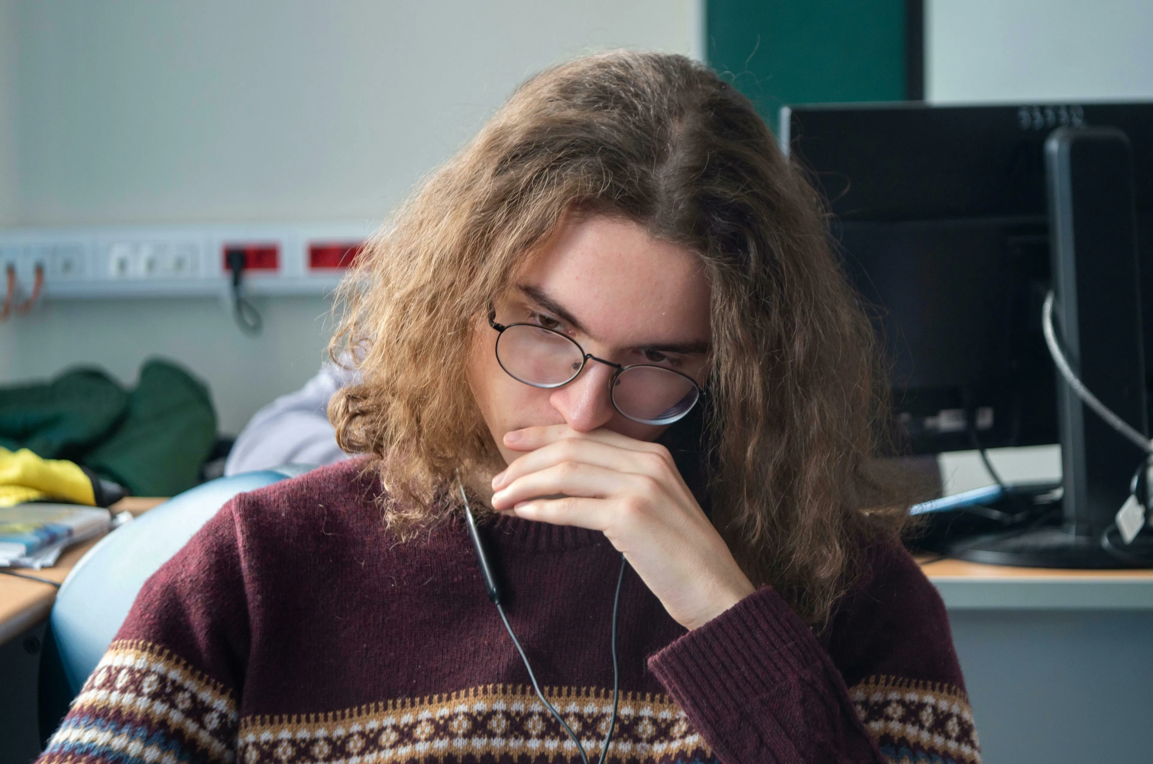 A thoughtful young man with long curly hair and glasses, wearing a patterned sweater, pondering how to handle being wrong.