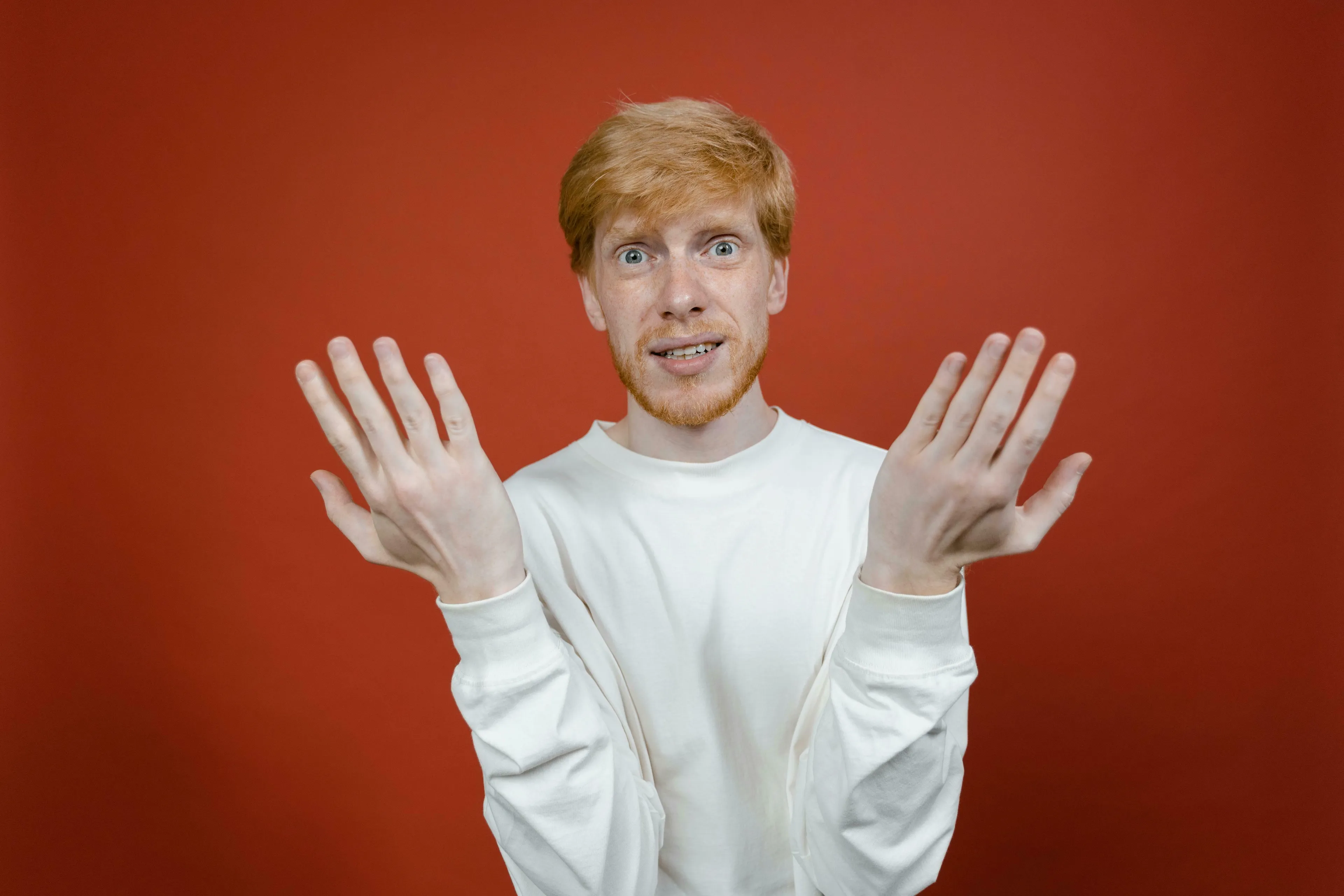 A confused redhead man in a white long-sleeve shirt shrugs, palms open. Represents handling being wrong.