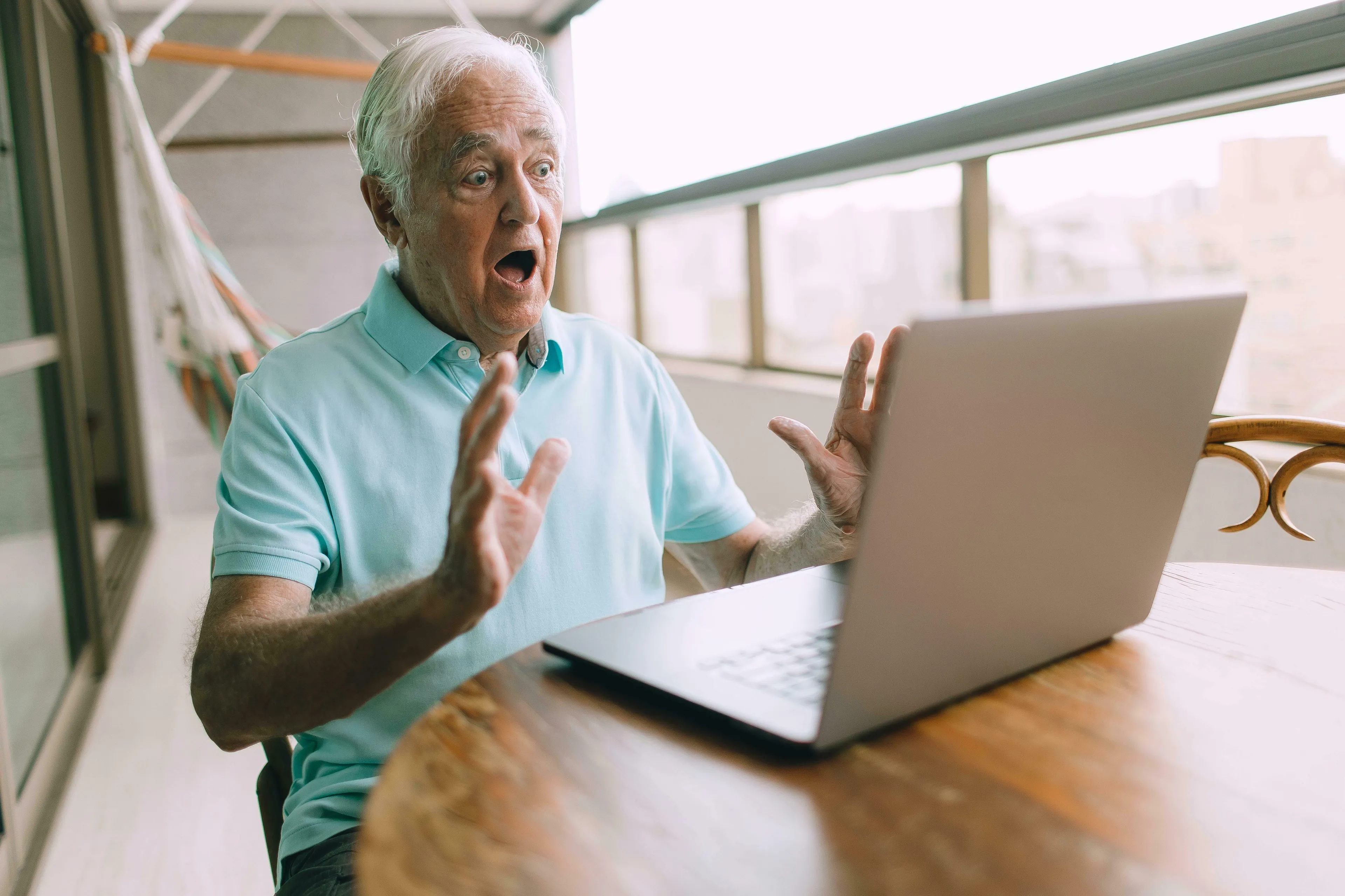 An older man in a light blue polo shirt looks shocked at his laptop, hands raised, demonstrating being wrong.