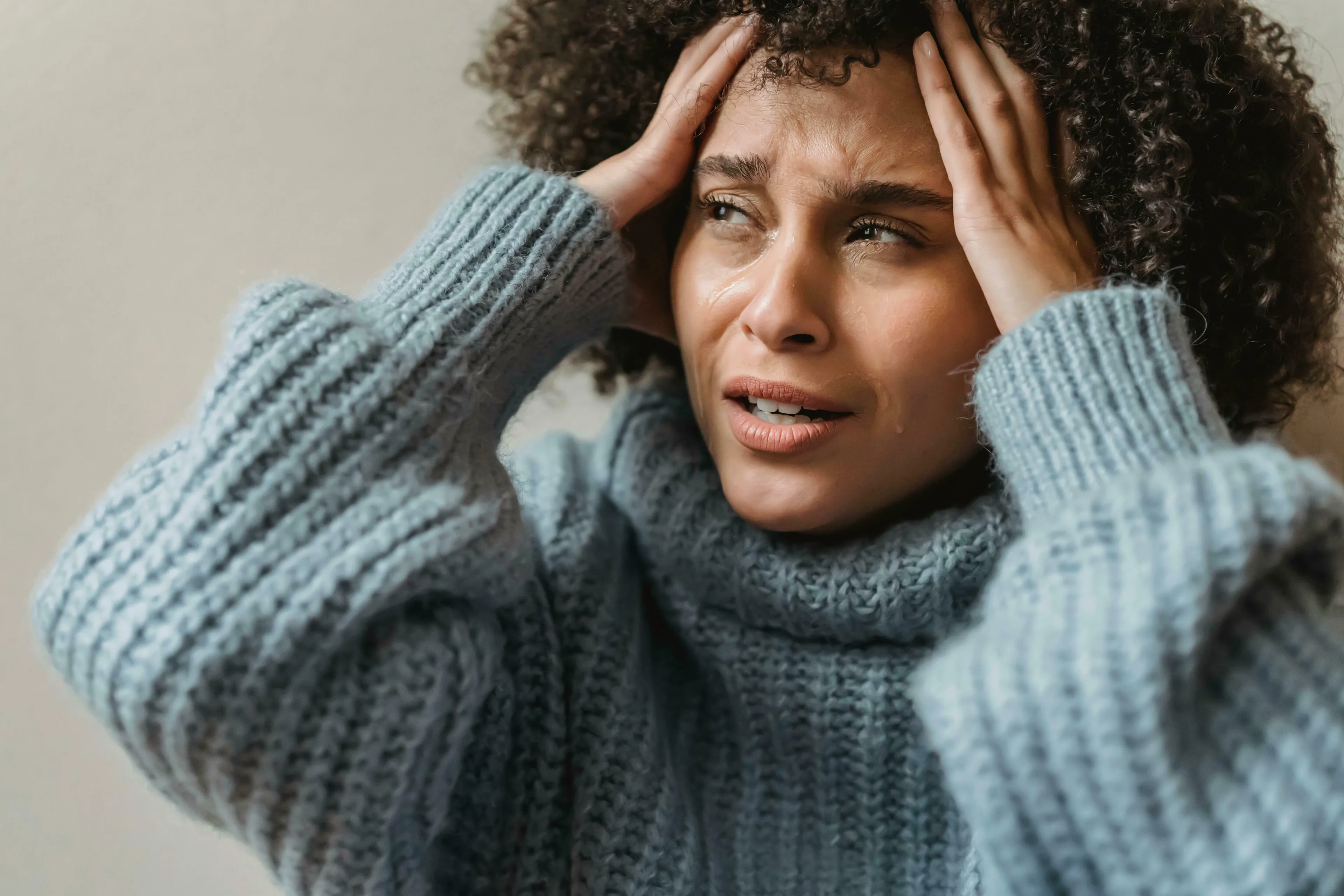 A woman with curly hair, wearing a blue knitted sweater, with tears on her cheeks and hands on her head, looking distressed, expressing her feelings when being wrong.