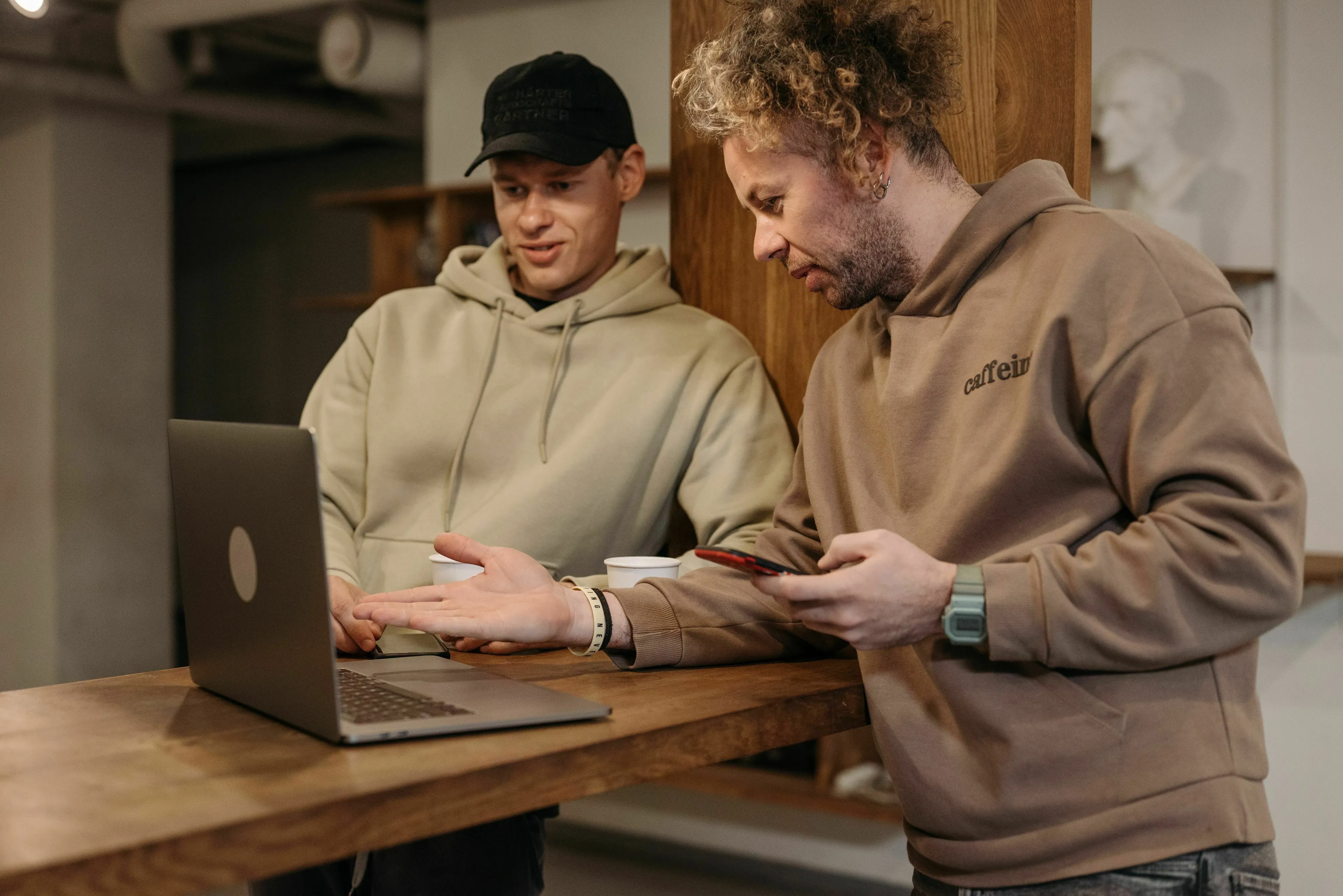 Two men, one in a cap, discussing something on a laptop. The man with curly hair holds a phone, appearing to handle being wrong.