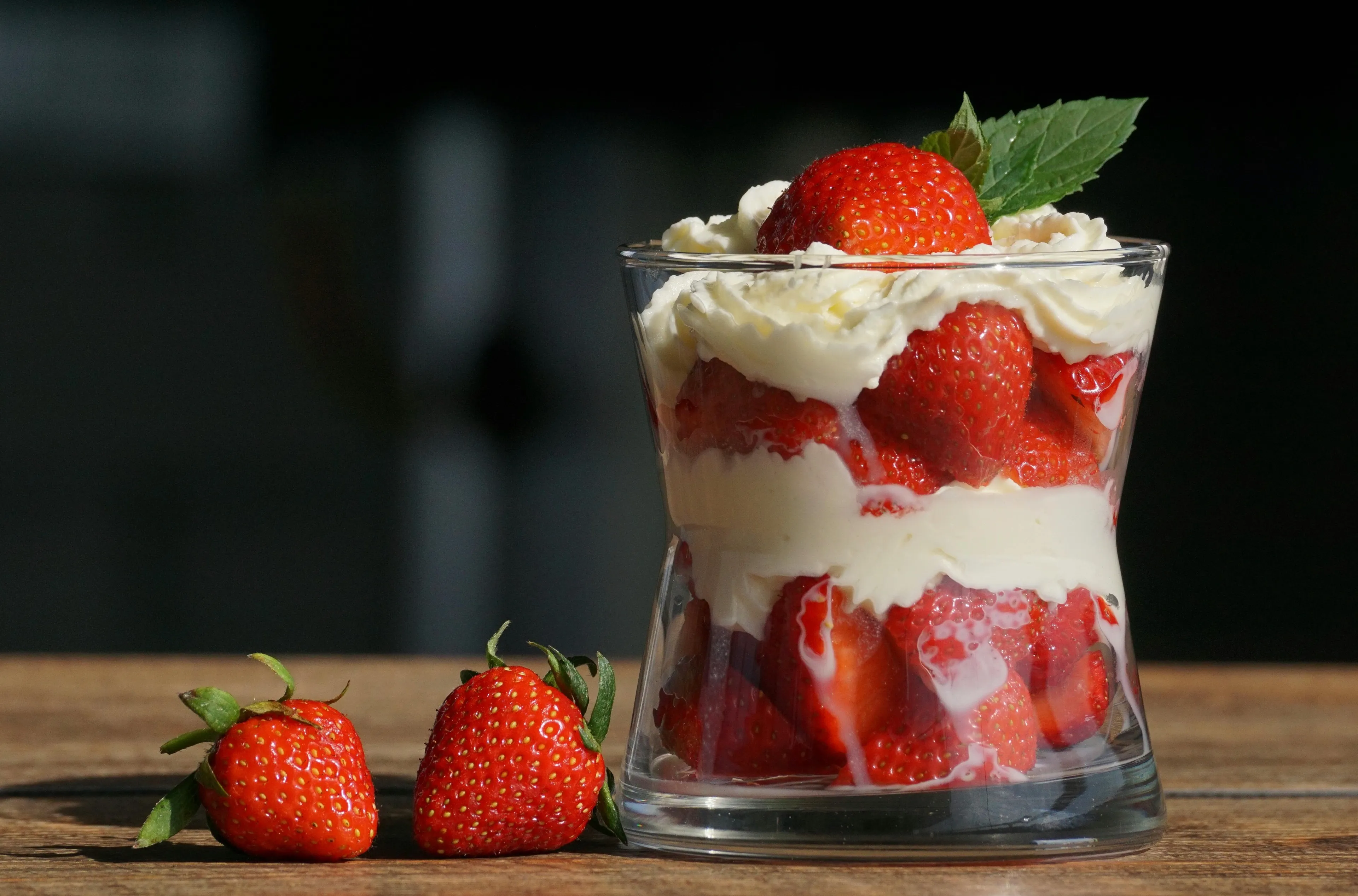A fresh strawberry trifle with whipped cream layers in a glass, alongside two whole strawberries on a wooden surface.