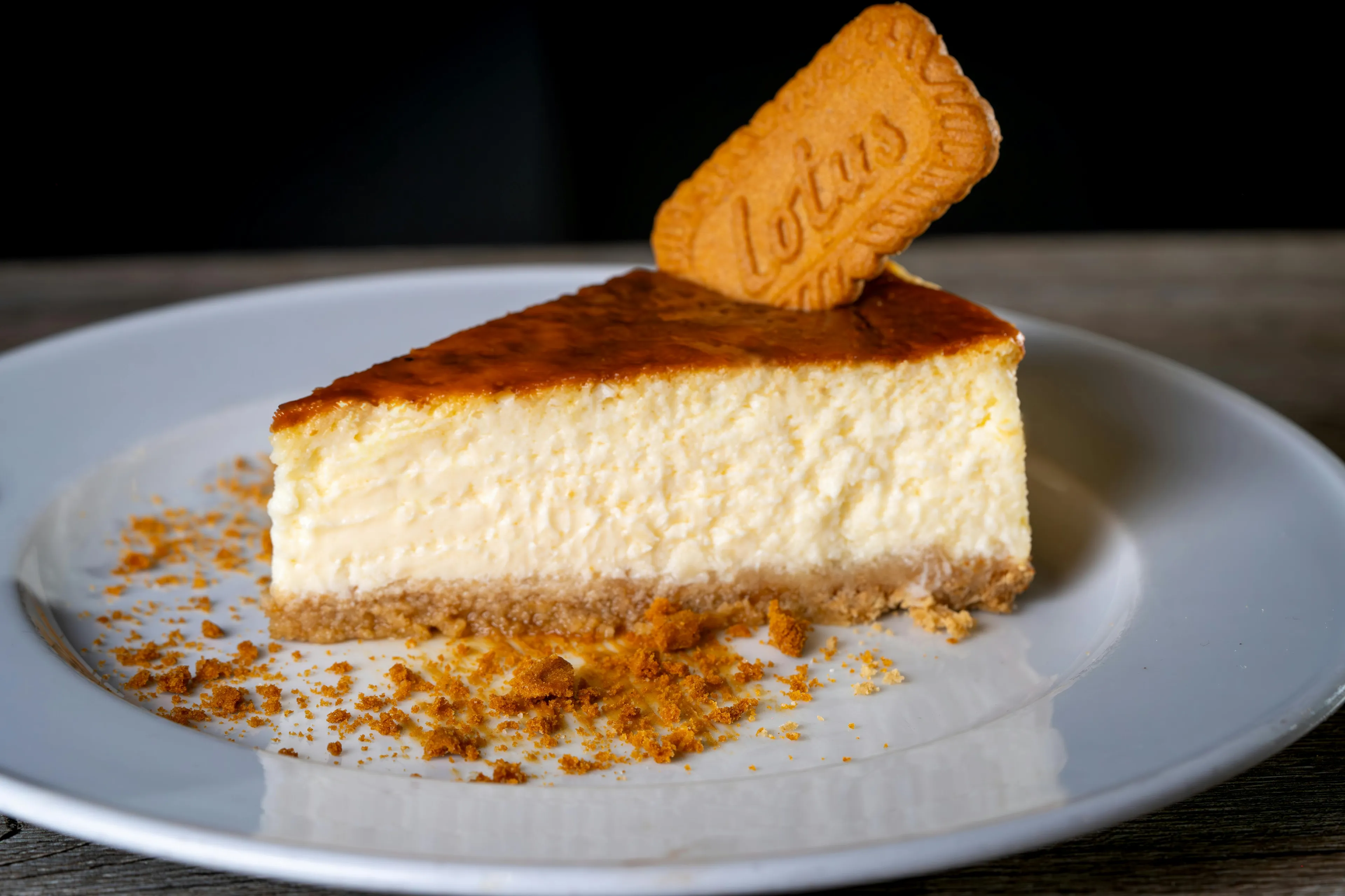 A slice of cheesecake on a white plate, topped with a Lotus Biscoff cookie and crumbled biscuits. A delicious dessert.