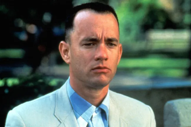 Forrest Gump, played by Tom Hanks, wears a light suit and blue checkered shirt, looking pensive.