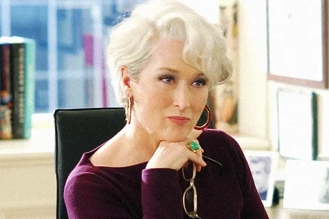 Miranda Priestly, with white hair, purple top, large gold earrings, holding glasses. Fictional character.