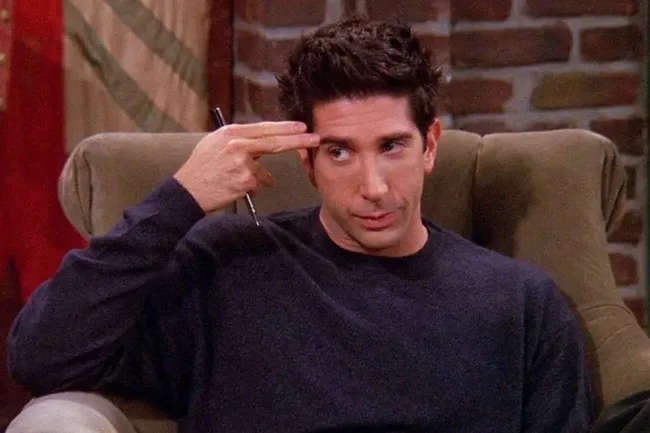 Ross Geller, in a blue sweater, sitting and making a hand gesture like a gun to his head, looking exasperated.