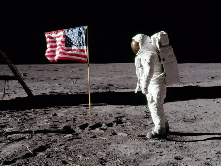 Astronaut in a white spacesuit next to an American flag planted on the lunar surface during the Moon Landing.