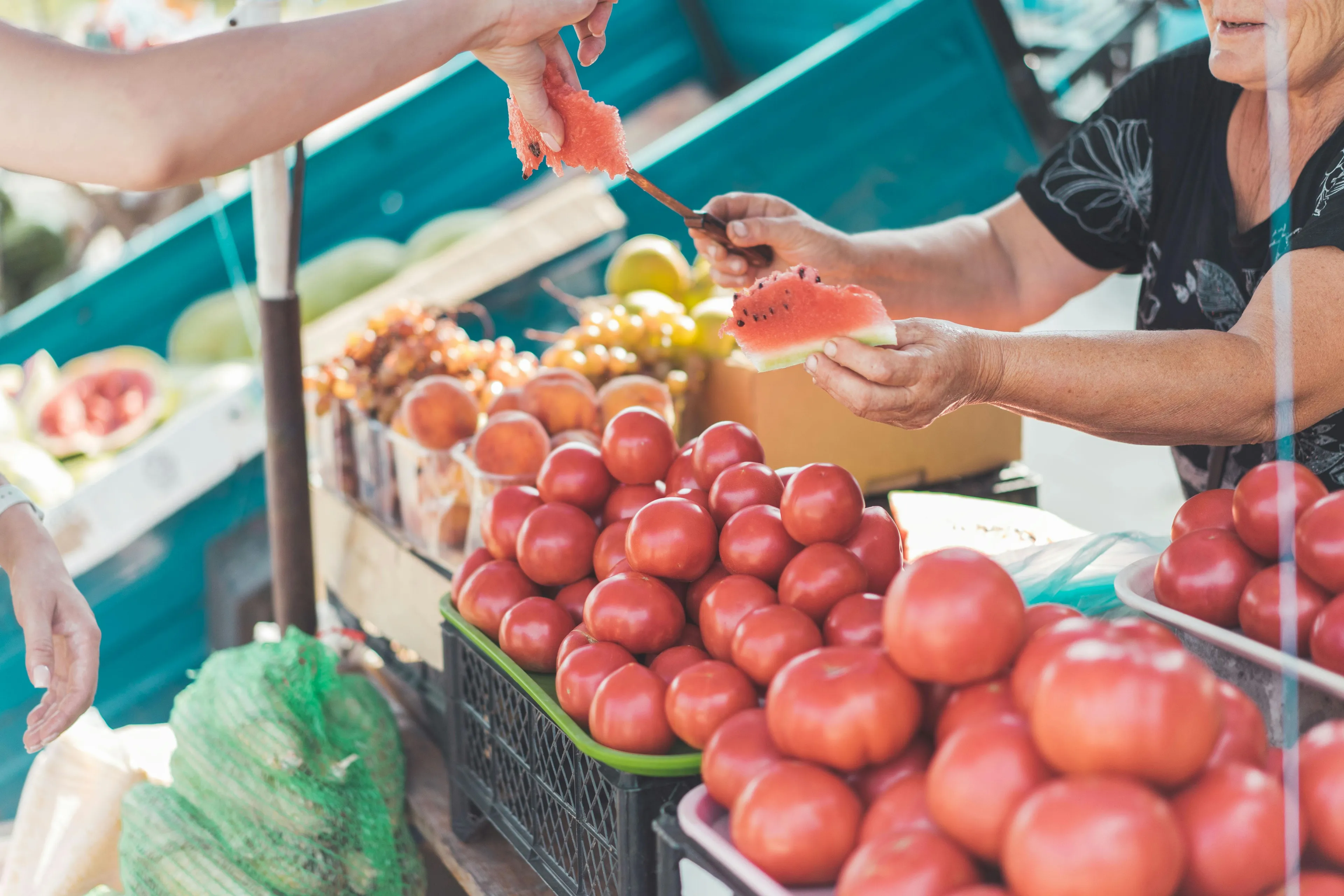 Hands exchange watermelon at a farmer's market, with vibrant red tomatoes and other fresh produce in the foreground.