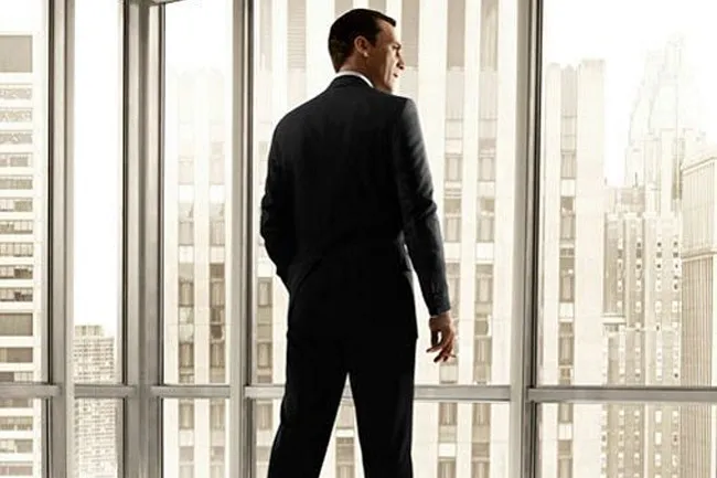 Don Draper from Mad Men in a dark suit, looking out a large window at a city skyline, holding a cigarette.
