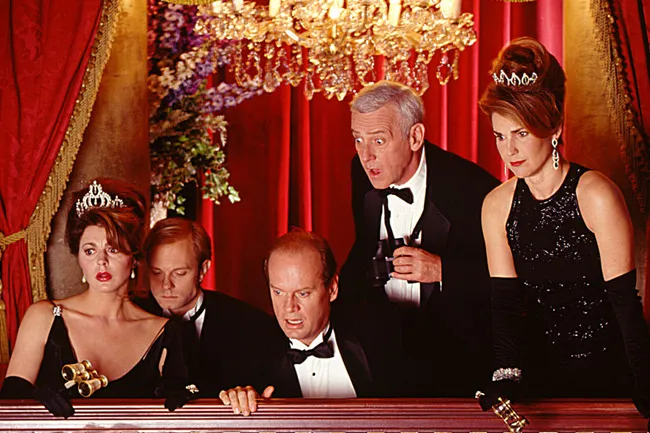 A surprised group from the TV show Frasier, dressed in formal attire, watches from a balcony at a performance.