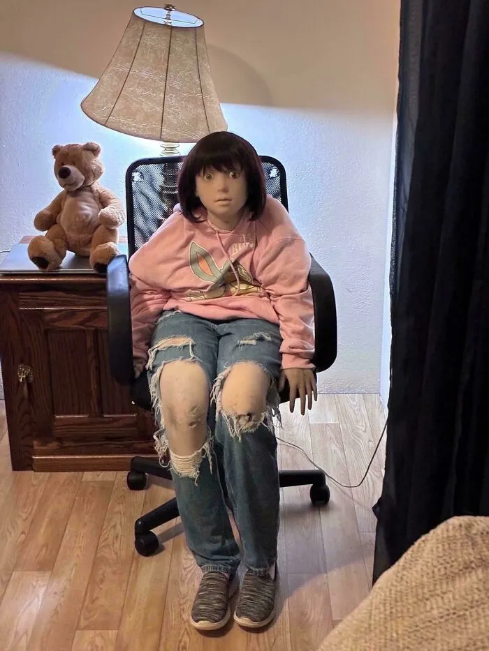 Creepy doll seated on a chair next to a teddy bear and lamp in a dimly lit room