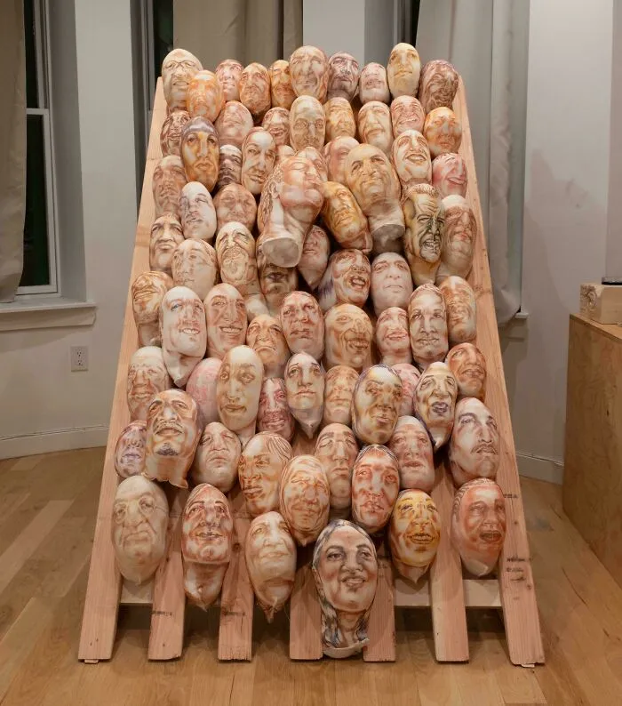 Art installation showing multiple carved faces on wooden objects arranged on a wooden rack