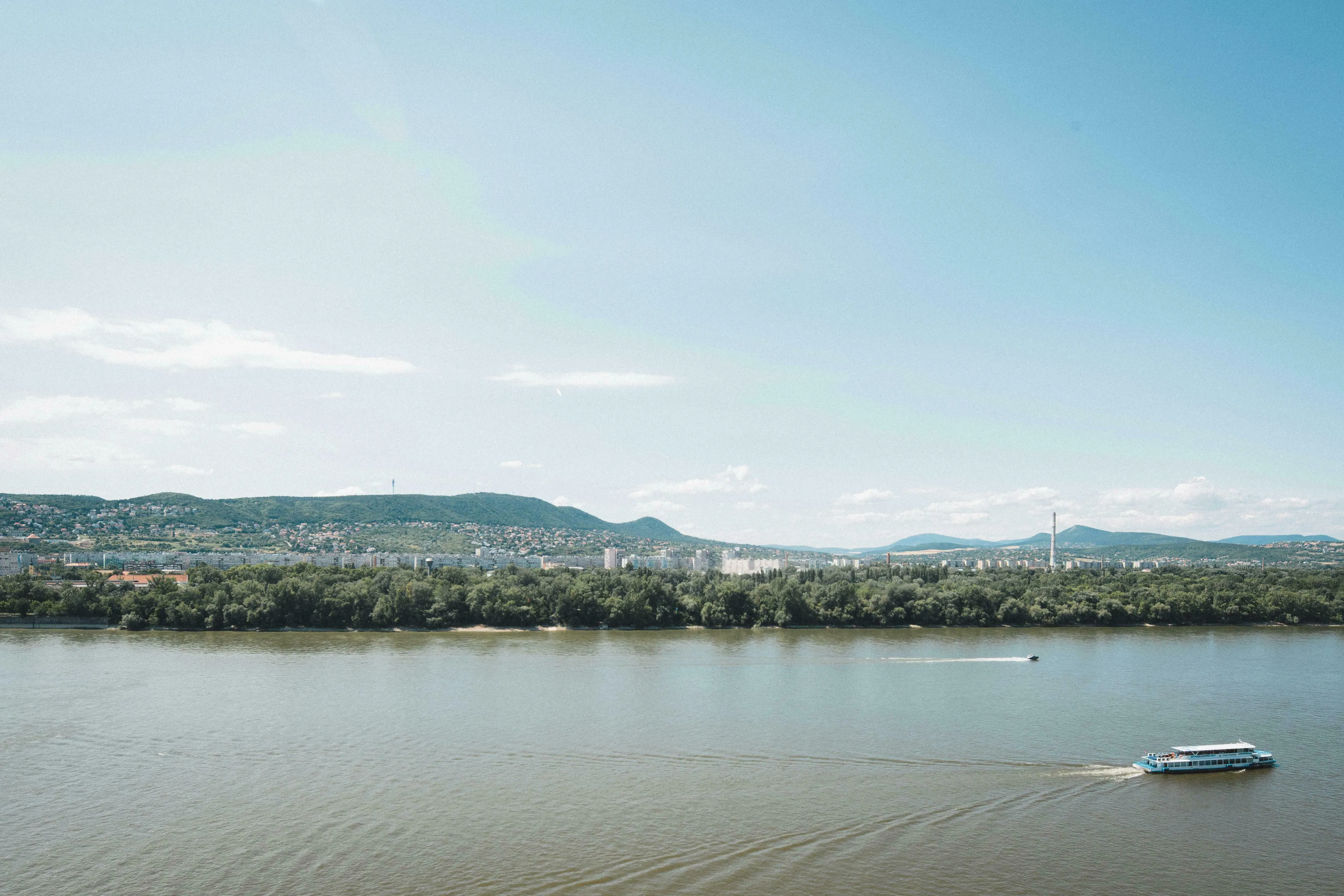 Panoramic view of the Danube River with boats, green banks, and a city against hills. A Pub Quiz question about the Danube.