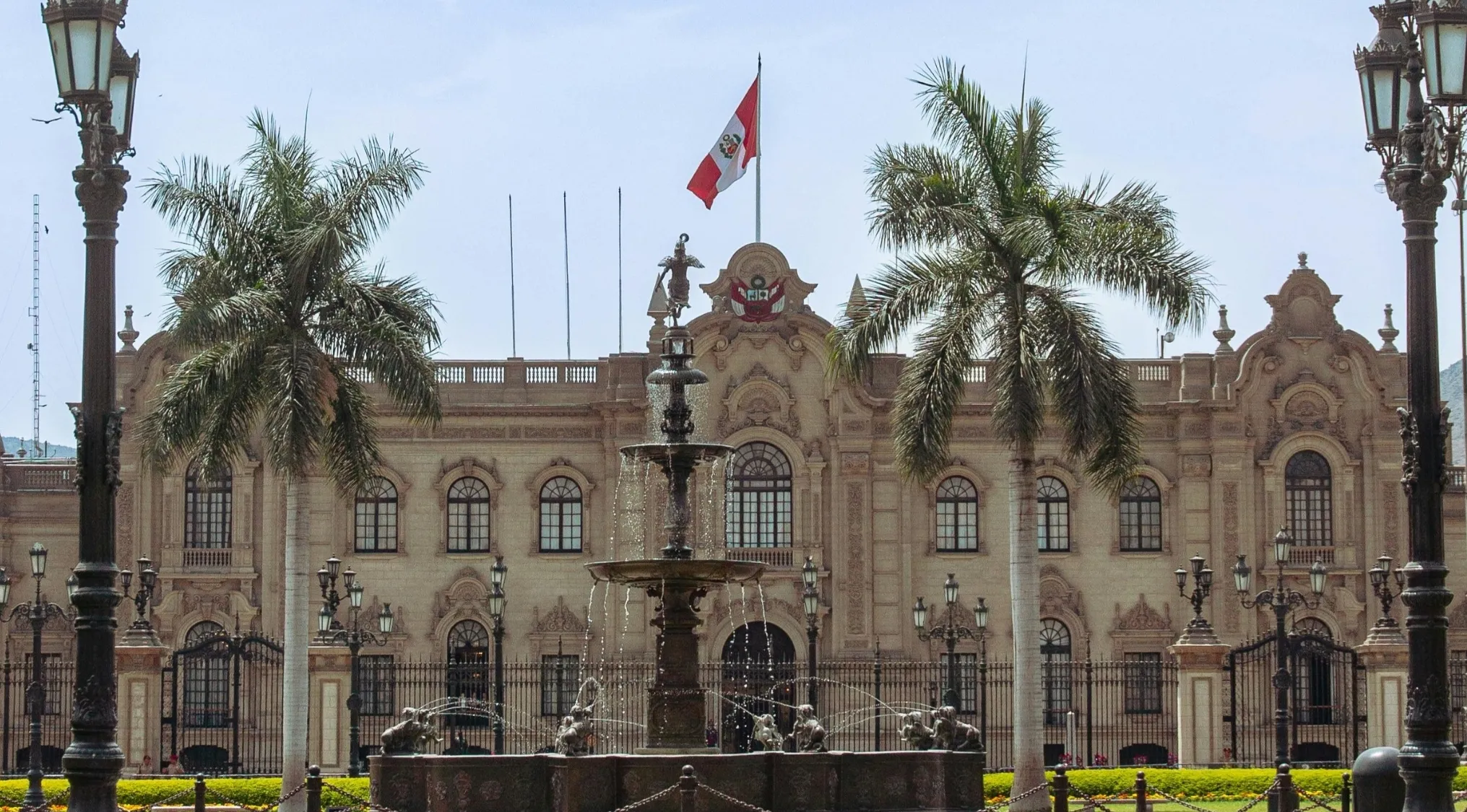 A grand stone building with a Peruvian flag, a fountain, and palm trees. Pub Quiz: Name the capital city of Peru.