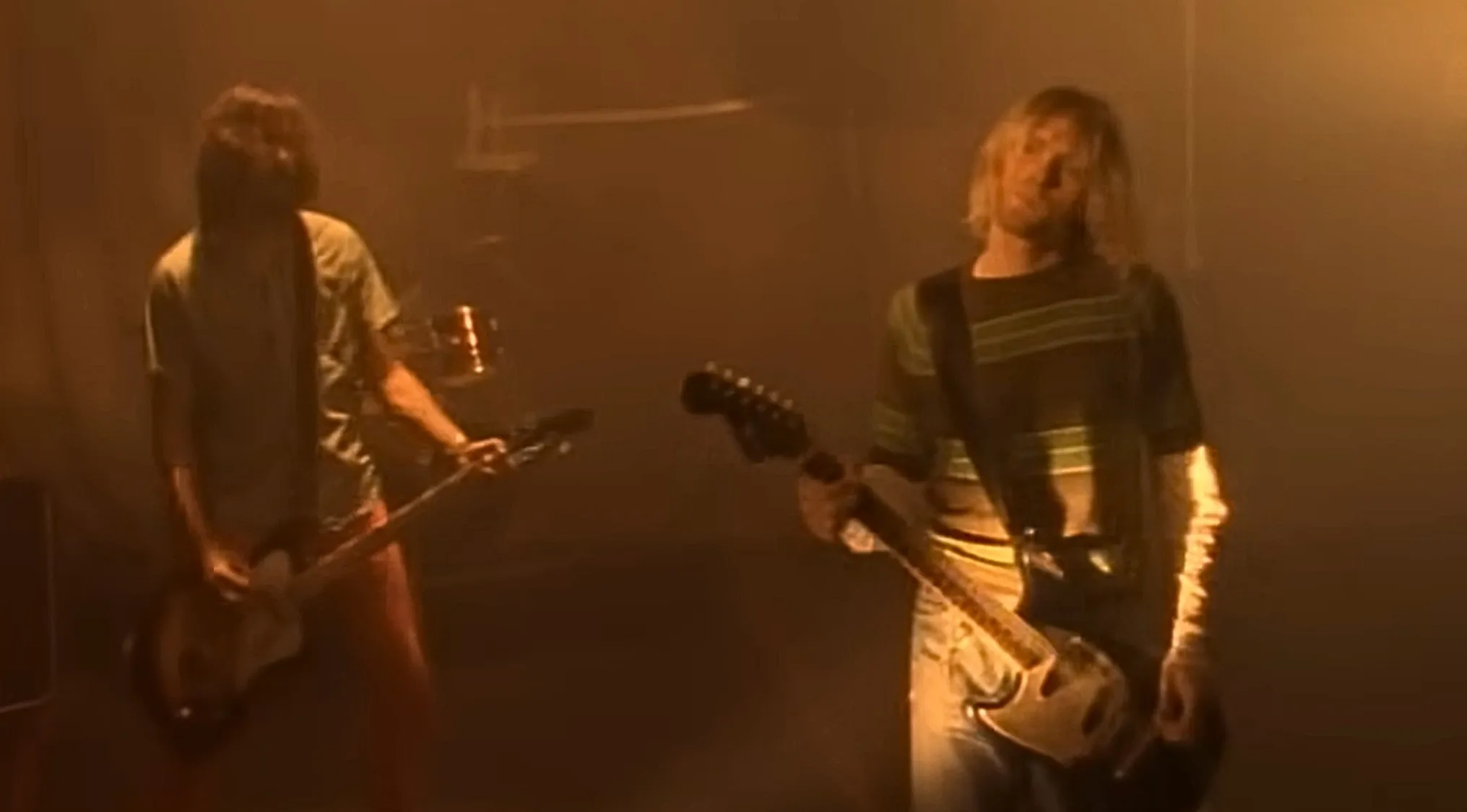 Two musicians in a smoky, dimly lit room, playing electric guitars. The frontman of Nirvana is on the right.