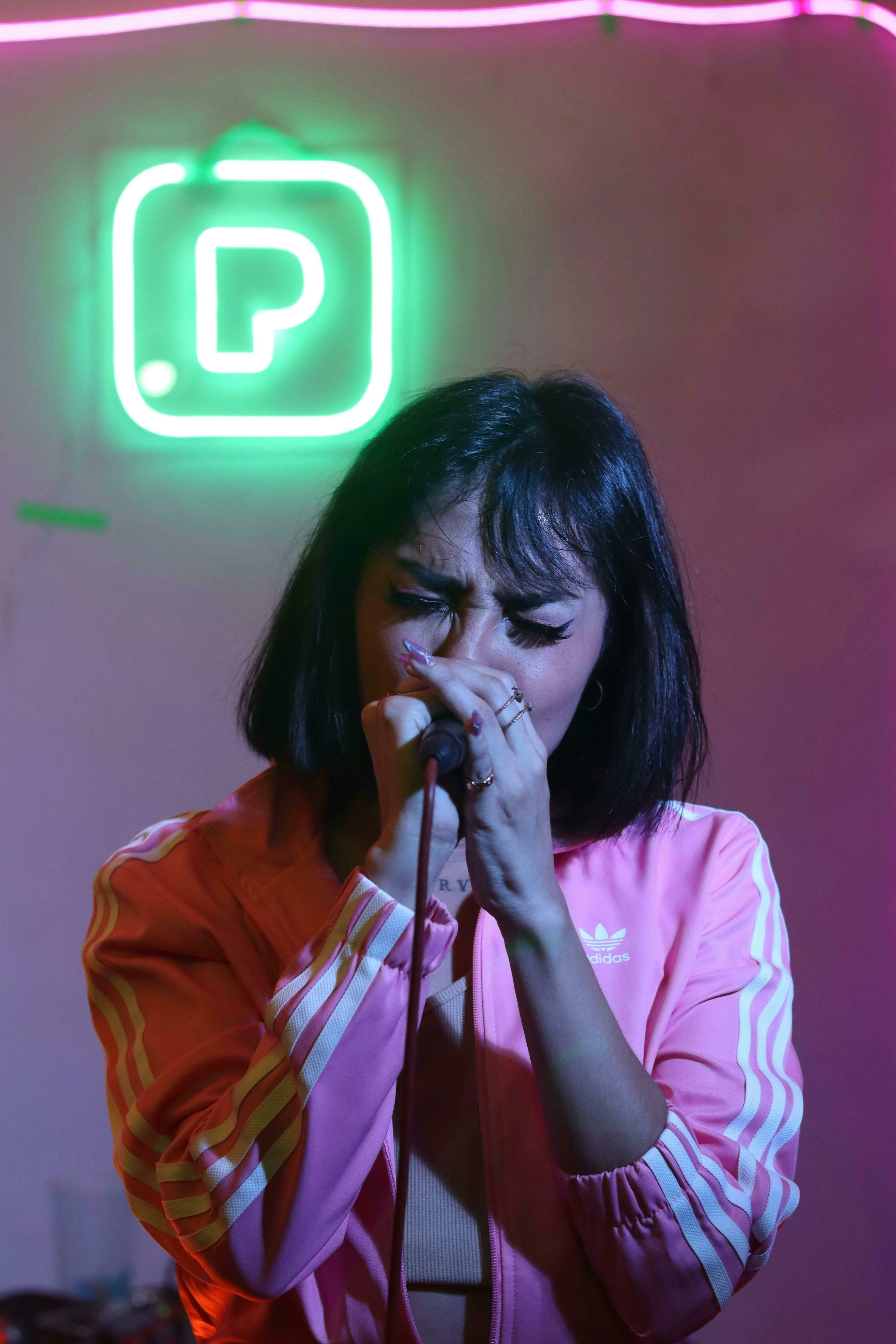 A woman in a pink tracksuit sings into a microphone during a pub quiz, with a green neon P in the background, thinking about where karaoke originated.