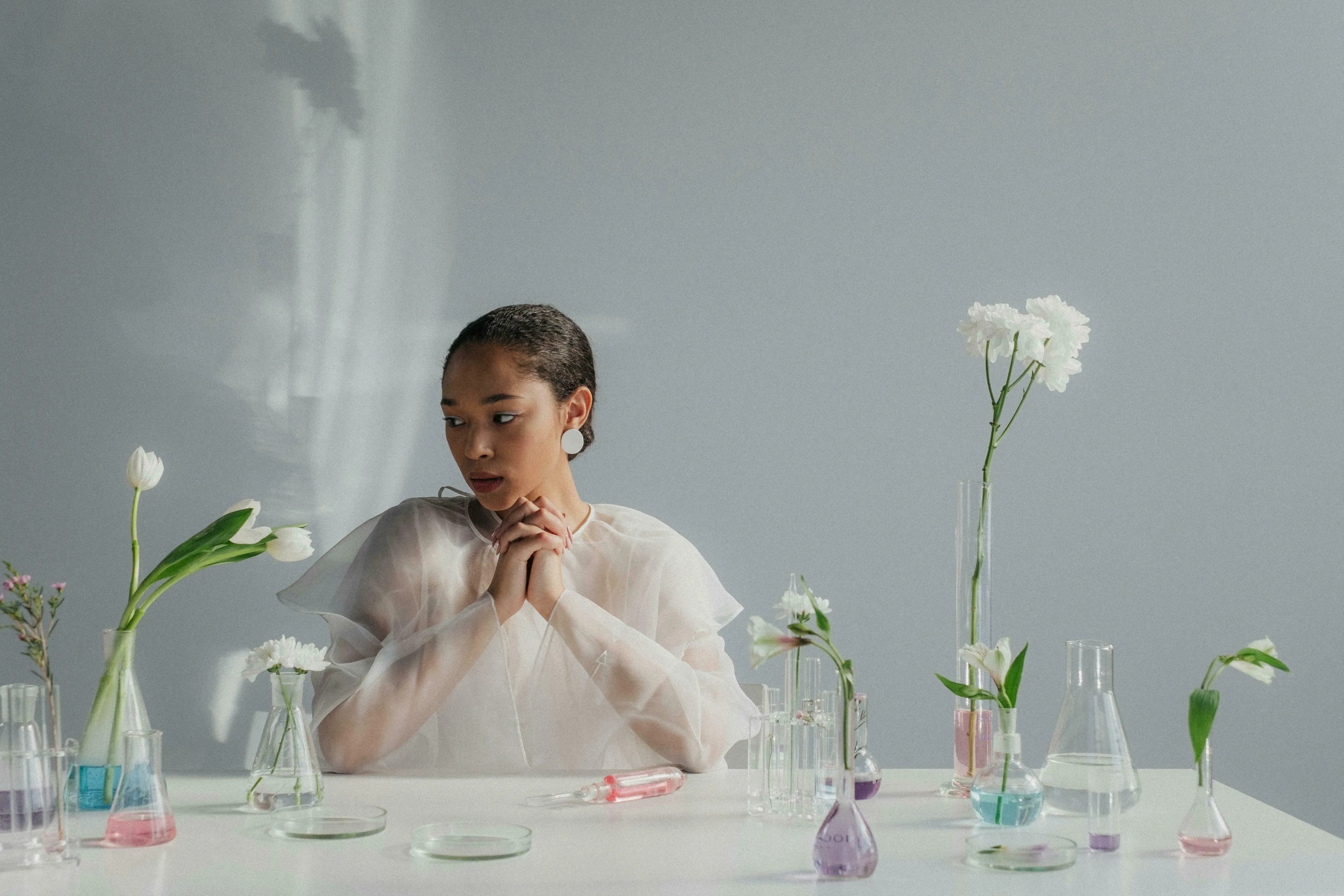 A young woman in white, surrounded by beakers and flowers, pondering a Pub Quiz question about female hormones.