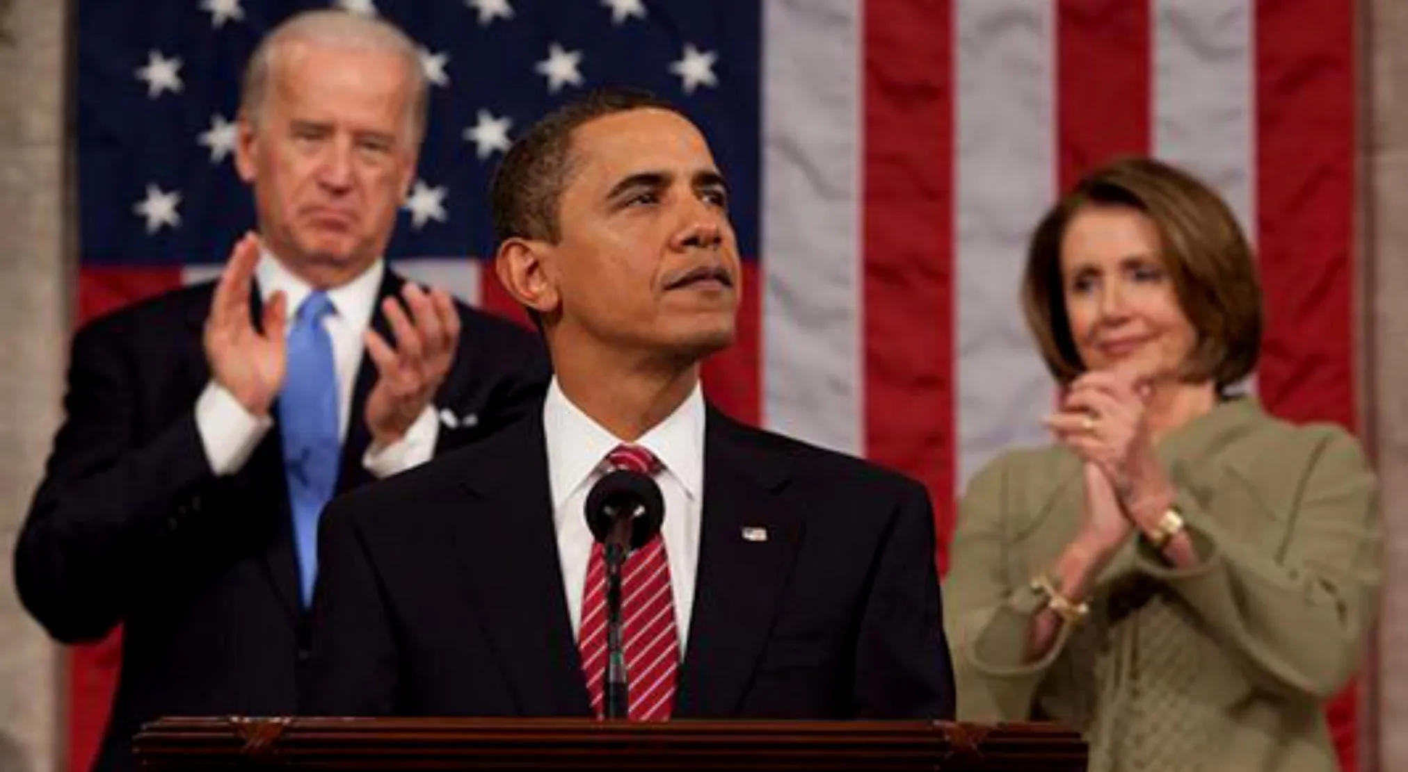 Barack Obama speaks at a podium, with Joe Biden and Nancy Pelosi clapping behind him, against a USA flag.