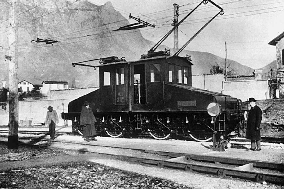 A black and white image of an early railroad train with three people near it. Railroads transformed travel during the Industrial Revolution.