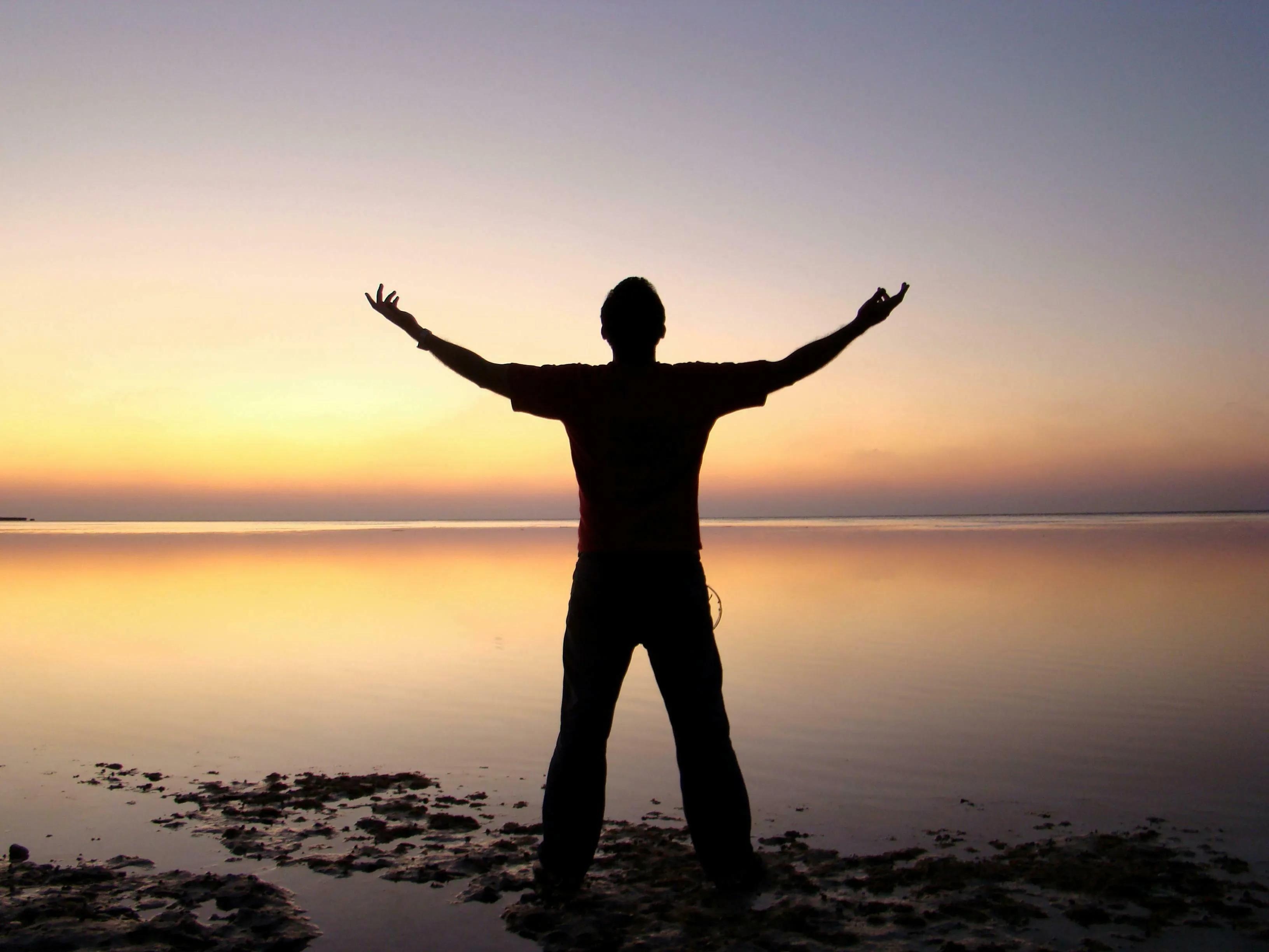 Silhouette of a person with outstretched arms facing a serene sunset over water. Reflects true nature and time of day.