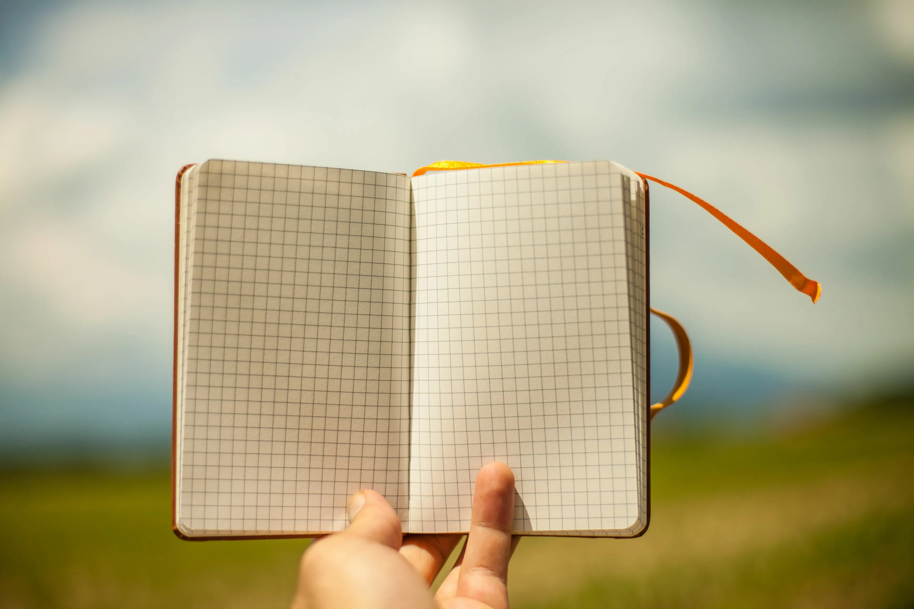 A hand holding an open, blank grid notebook with an orange bookmark, suggesting a fresh start for writing or drawing. Blank notebook for your true nature.