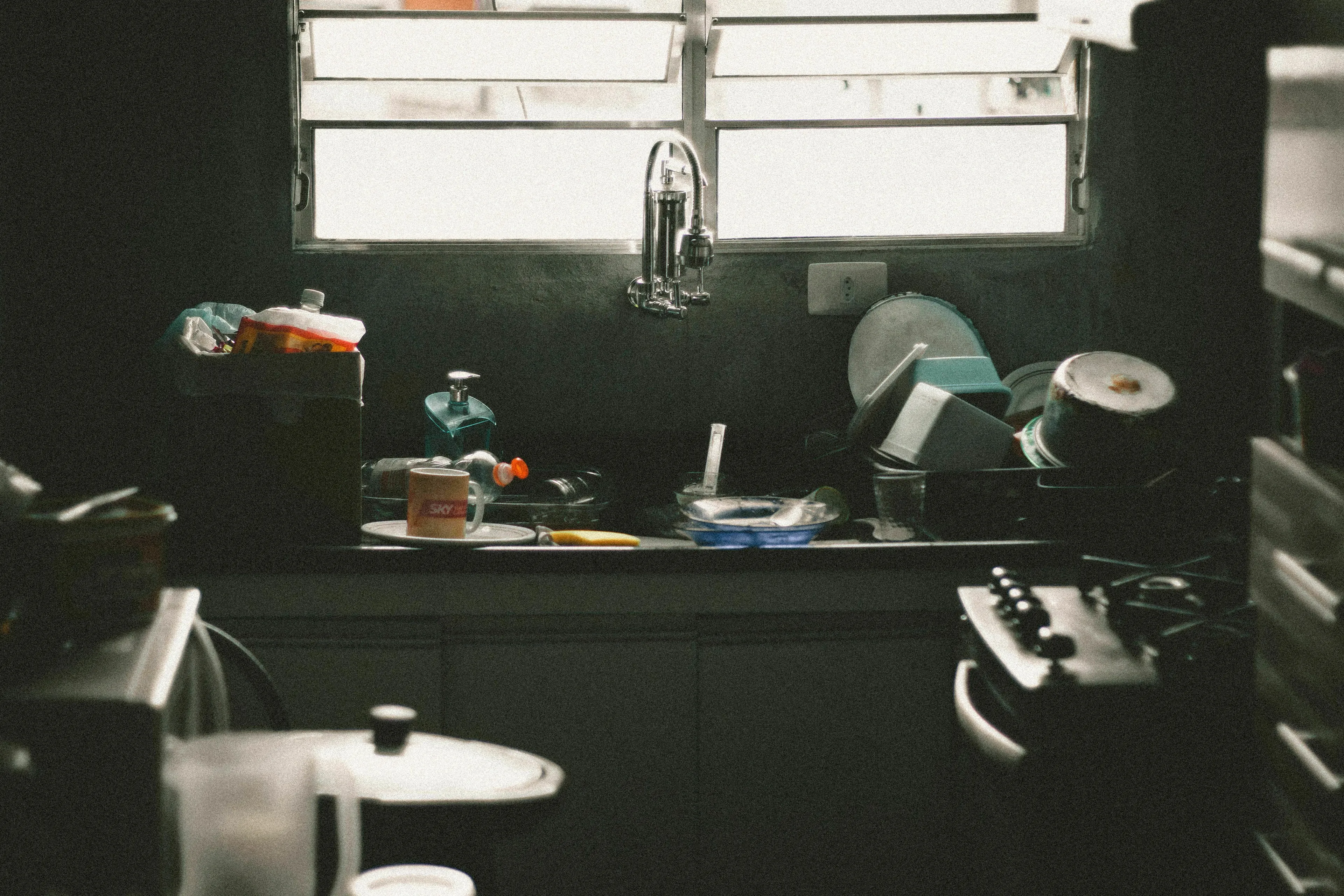 A messy kitchen sink area with dirty dishes and dim lighting. Which Season, kitchen is a mess, true nature.