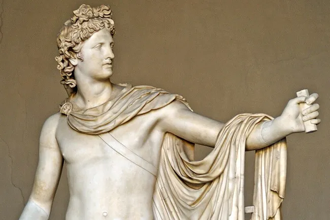 A marble statue of Apollo, wearing a laurel wreath and draped toga, looking to the right. He holds an object in his hand.