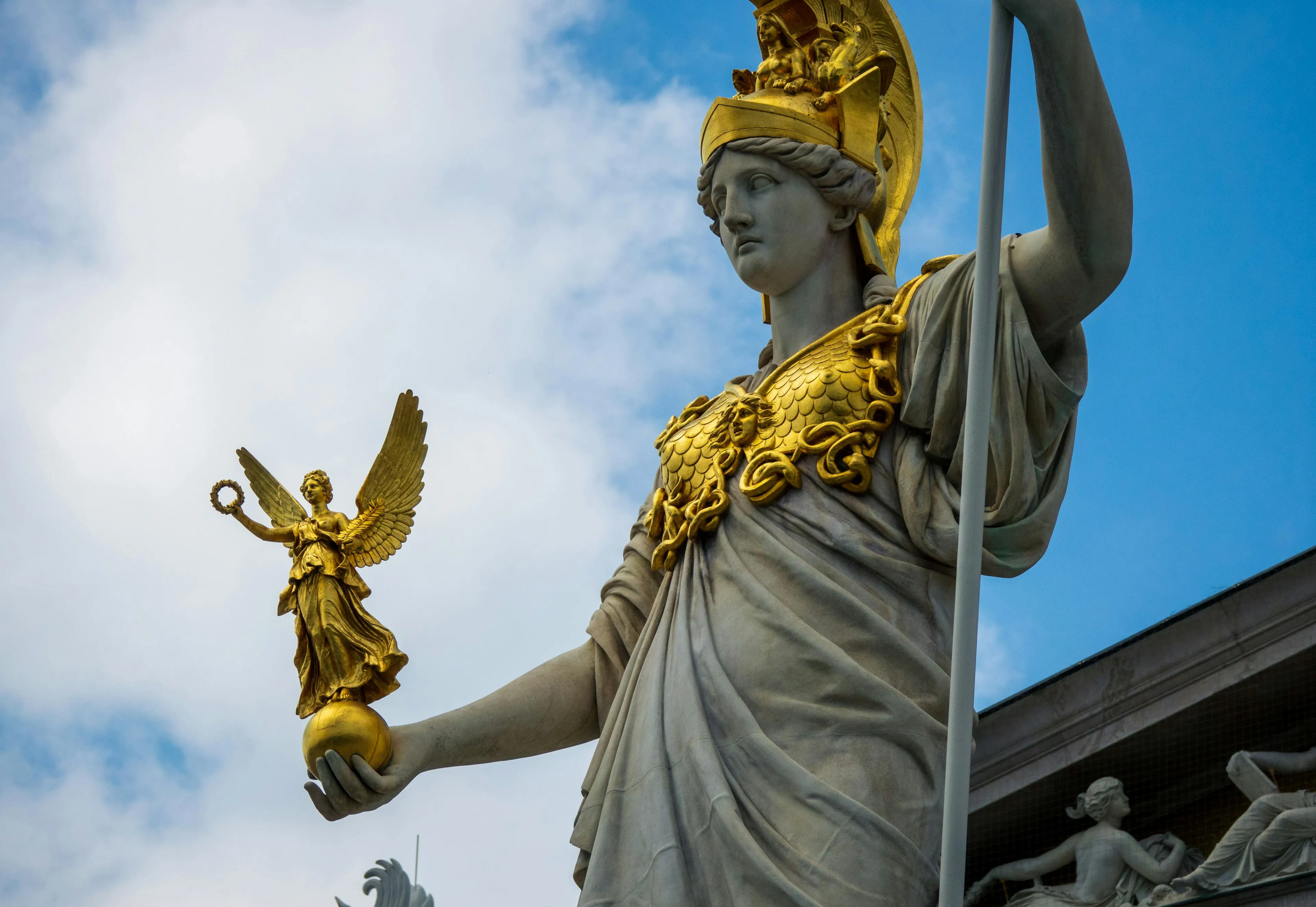 A majestic Athena statue in white and gold, holding a golden winged figure, set against a blue, cloudy sky.