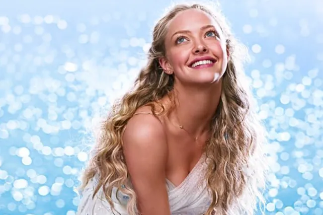 Amanda Seyfried in Mamma Mia, smiling with long wavy blonde hair, looking up against a sparkling blue background. Movie to watch on repeat.