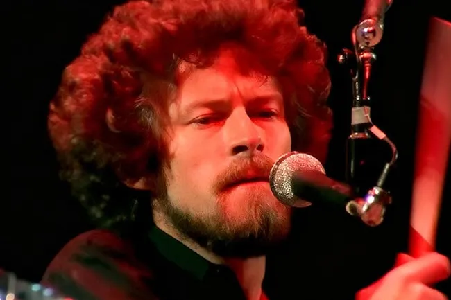 A man with curly red hair and a beard, singing into a microphone, illuminated by red stage lights. Inner world song.