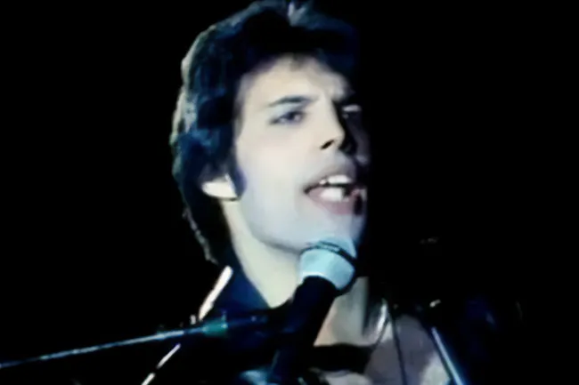 Freddie Mercury singing Don't Stop Me Now with Queen, eyes open, mouth open at a microphone on a dark stage.
