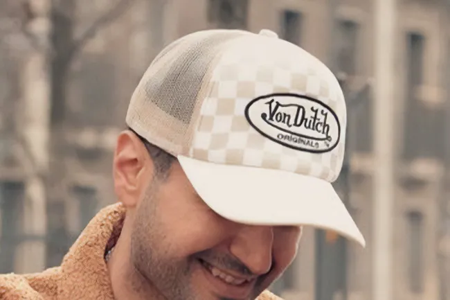 A smiling person wearing a cream and tan checkered Von Dutch hat, bringing back a classic fashion trend.