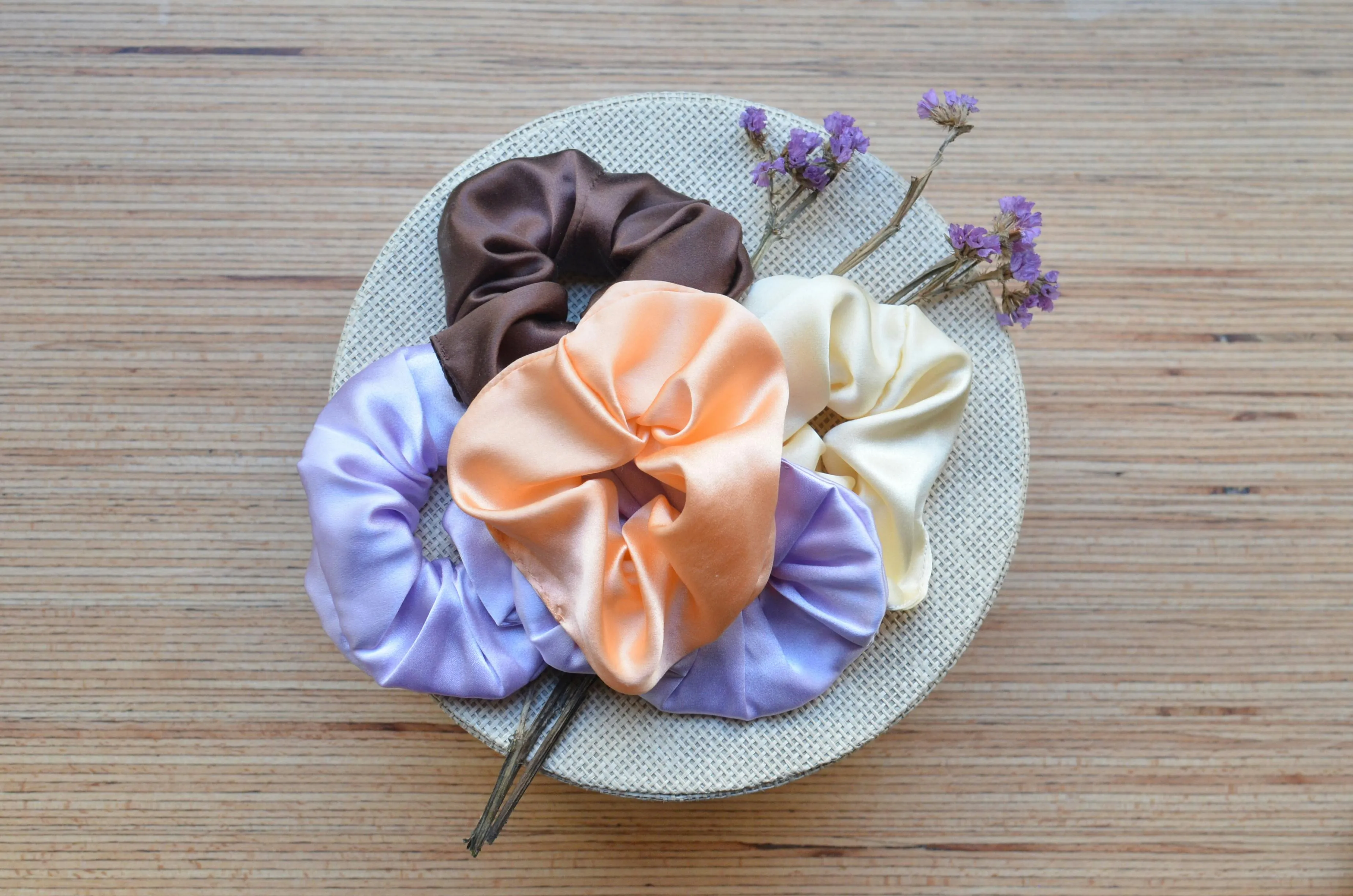A collection of colorful scrunchies in brown, peach, cream, and lavender on a round woven mat with dried purple flowers.